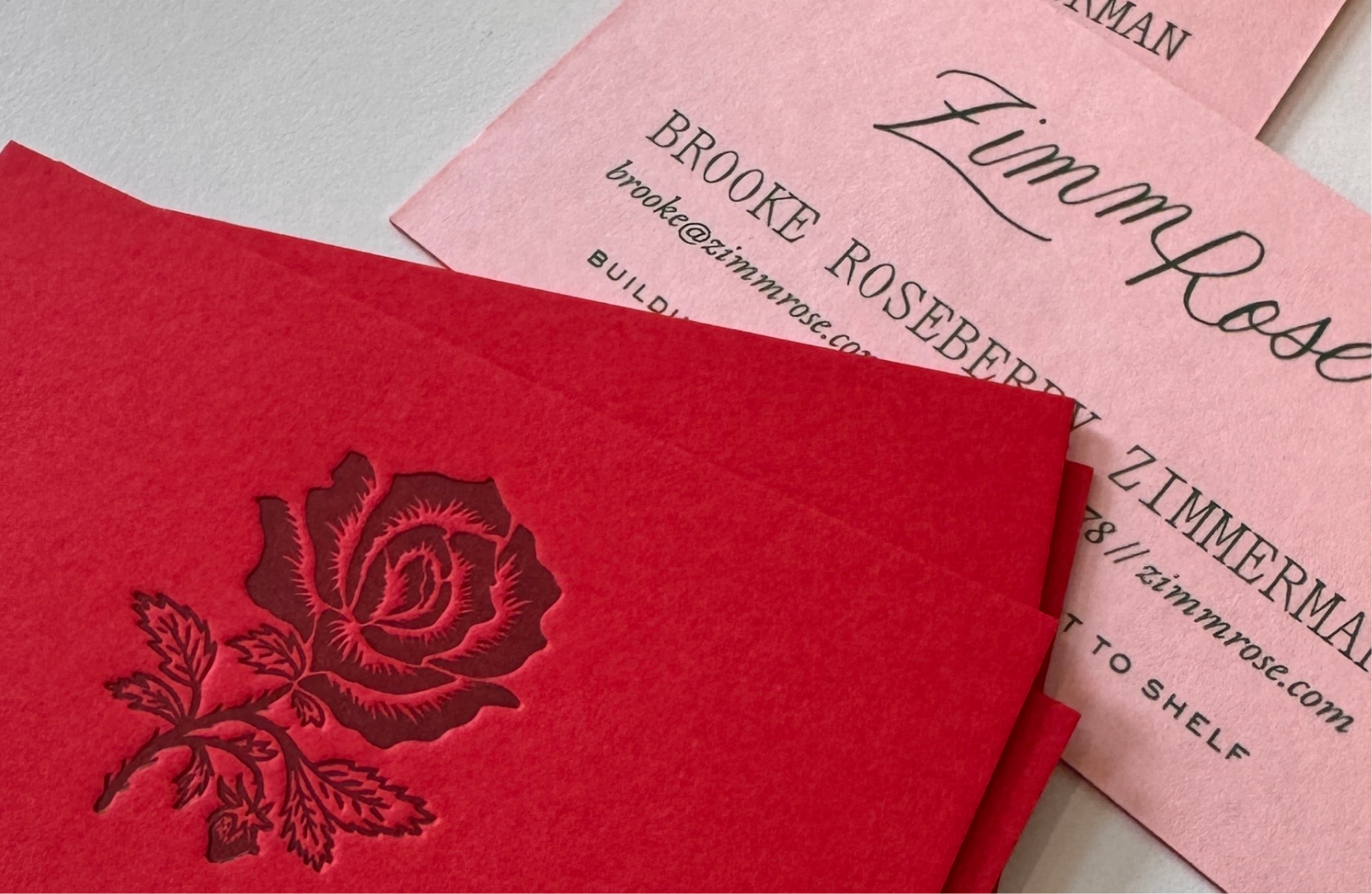 A close-up of red embossed rose cards layered with pink ZimmRose business cards featuring Brooke Roseberry Zimmerman’s contact details and refined branding elements.
