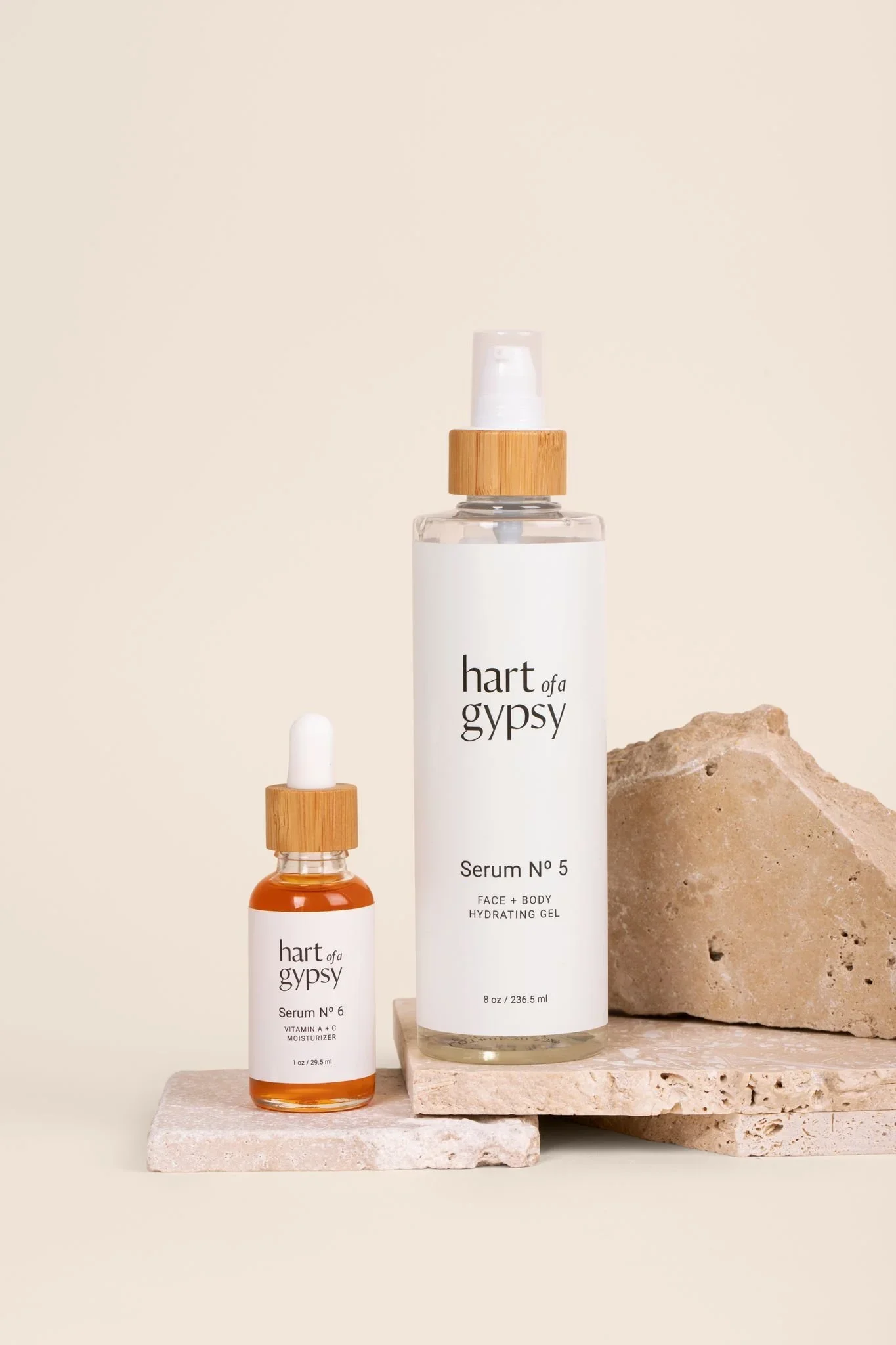 Two skincare products on beige stone slabs: an amber dropper bottle of Serum No. 6 and a clear pump bottle of Serum No. 5, both labeled “hart of a gypsy.”