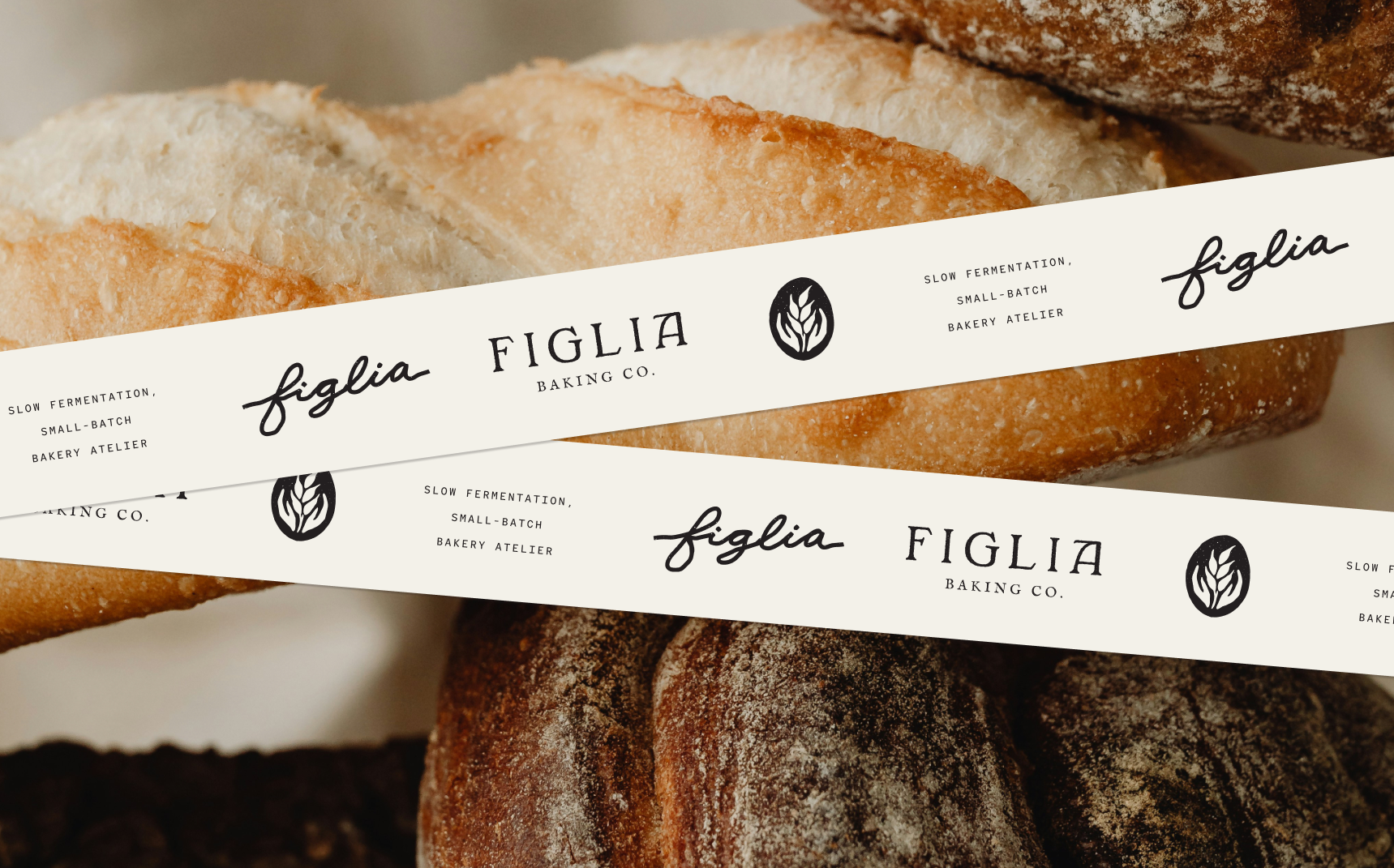 Several artisanal bread loaves arranged together with two cream-colored branded paper bands laid across them. The bands feature the Figlia Baking Co. logo, a stylized wheat emblem, and the text “Slow fermentation, small-batch bakery atelier.”
