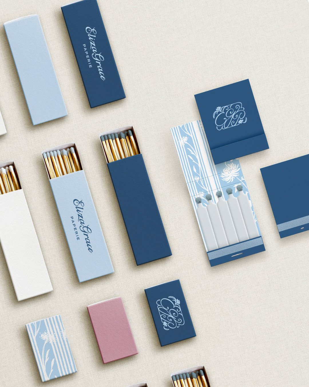An assortment of matchboxes and matchbooks in shades of blue, white, and pink arranged on a beige surface, each featuring elegant ElizaGrace Paperie branding and monogram logos, with some boxes open to reveal wooden matches.
