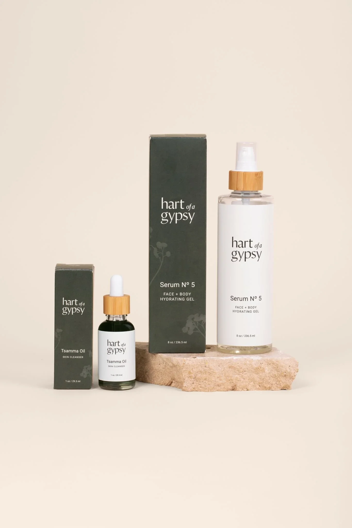 Two skincare products displayed on beige stone blocks: Tsamma Oil Skin Cleanser in a small dropper bottle and Serum No. 5 Hydrating Gel in a larger spray bottle, both with matching dark green boxes.