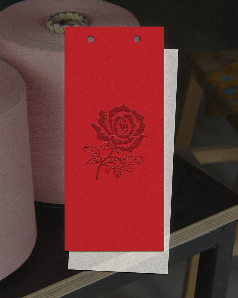 A red card with an embossed rose design layered over a white card, placed in front of two large pink thread spools, blending textile production with brand identity.