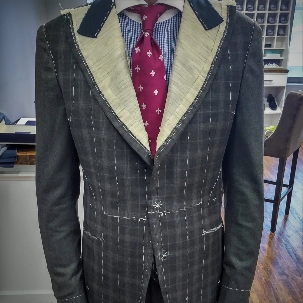 Partially constructed tailored suit on a mannequin, showing exposed stitching and unfinished lapels, with a red patterned tie and a blue checkered dress shirt underneath.