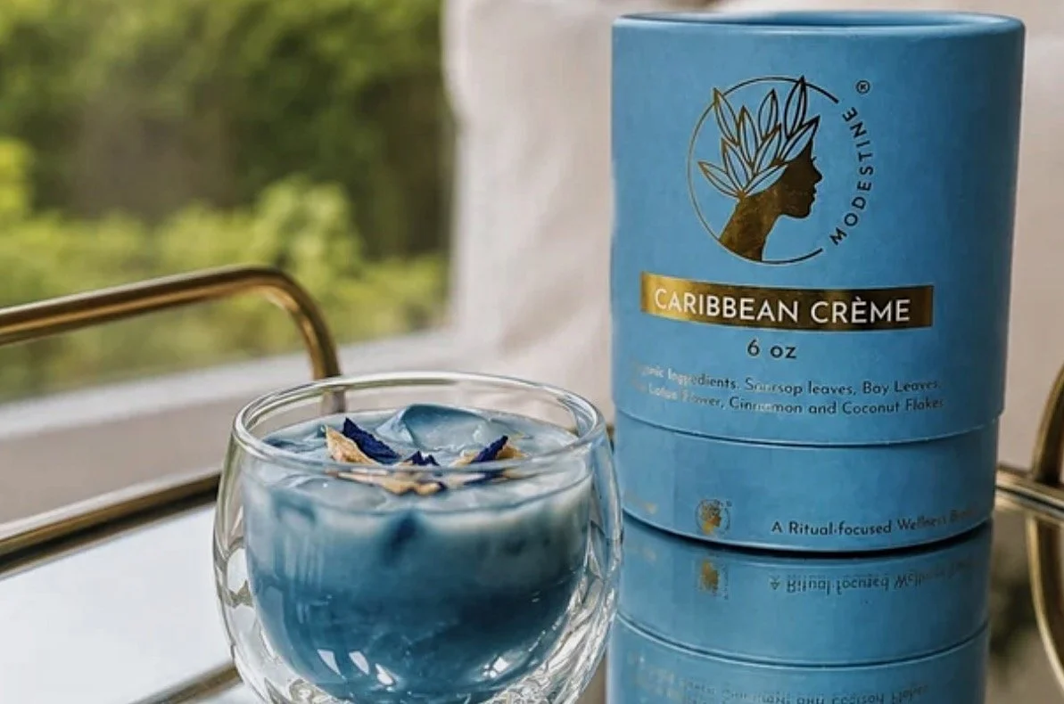 byModestine Caribbean Creme tea tin beside a vibrant blue iced tea cocktail on a mirrored tray with a tropical garden view.