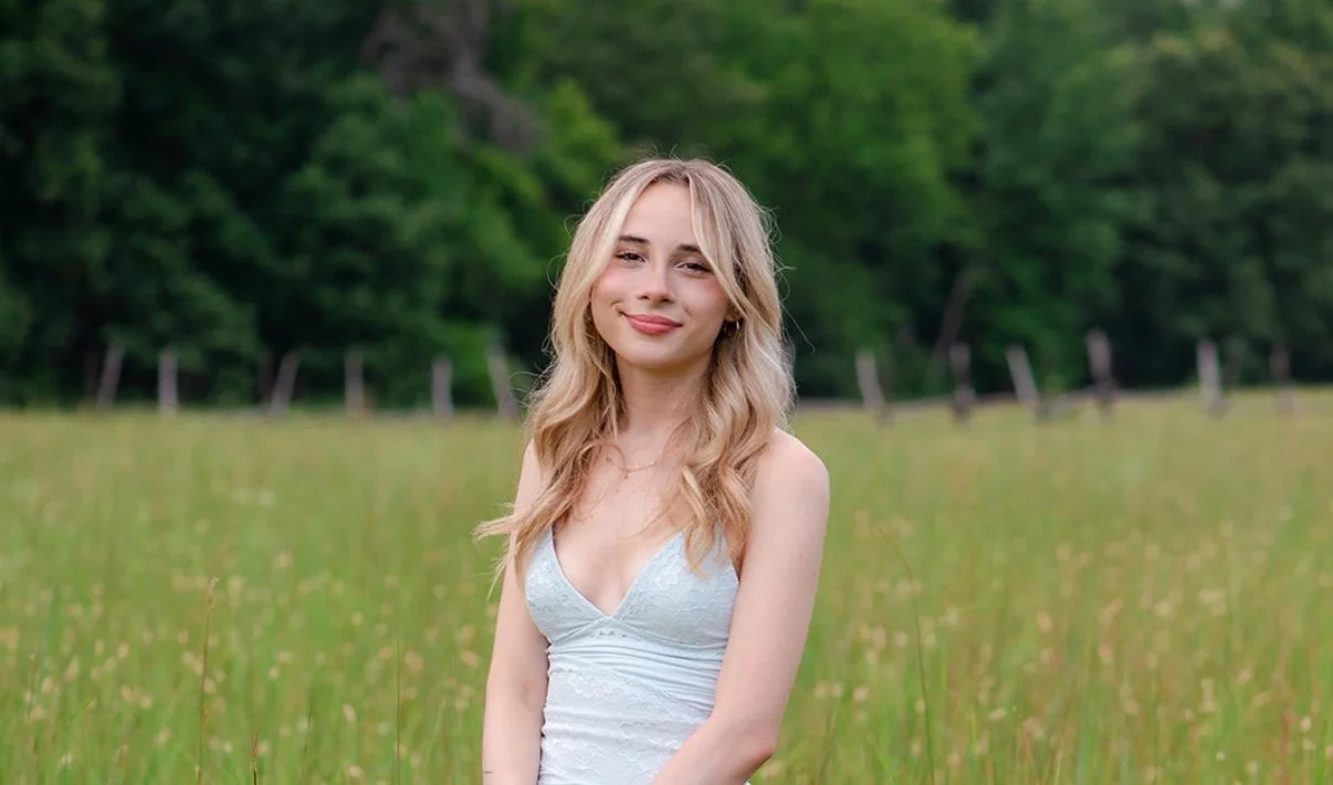 Young woman with long blonde hair smiling in a white dress standing in a grassy field with trees in the background.