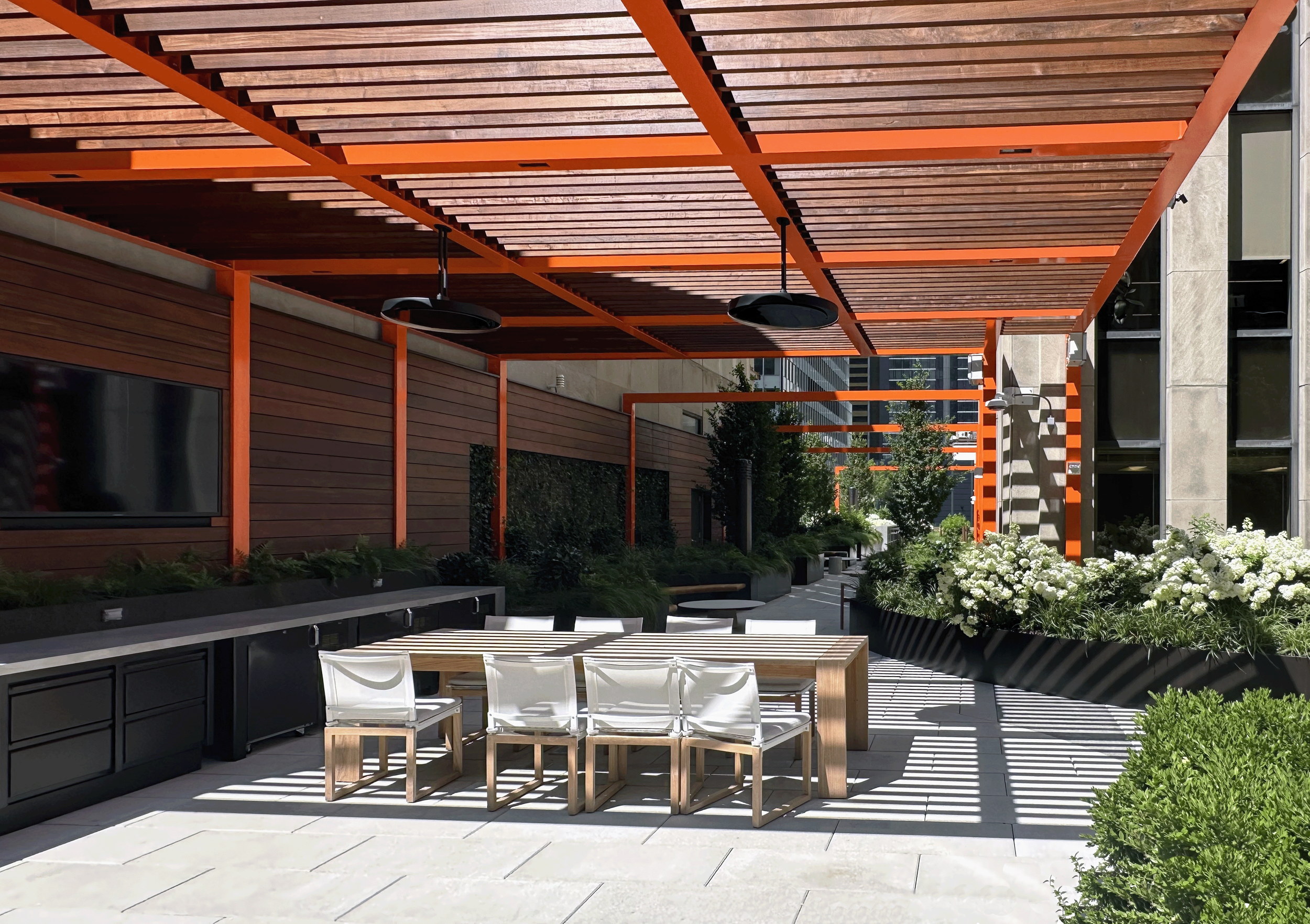 Outdoor patio with wooden pergola, white chairs, dining table, lush greenery, and flowering plants.
