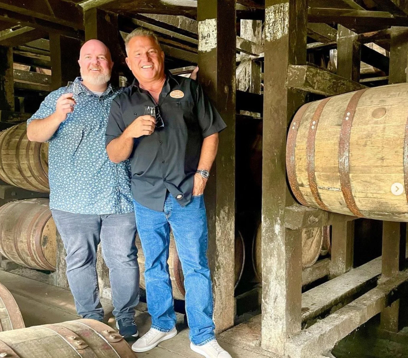 Two men standing inside a bourbon barrel cellar, holding glasses of dark liquor, smiling for the camera. There are wooden barrels stacked behind them.