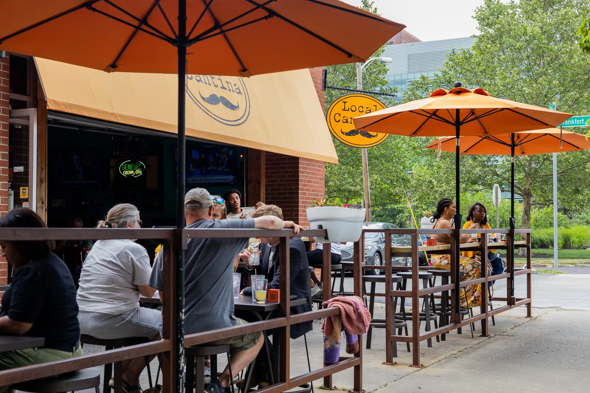 People dining outdoors at a restaurant with orange umbrellas. The restaurant has a sign that says 'Local Cantina' with a mustache graphic. The scene includes trees and modern buildings in the background.