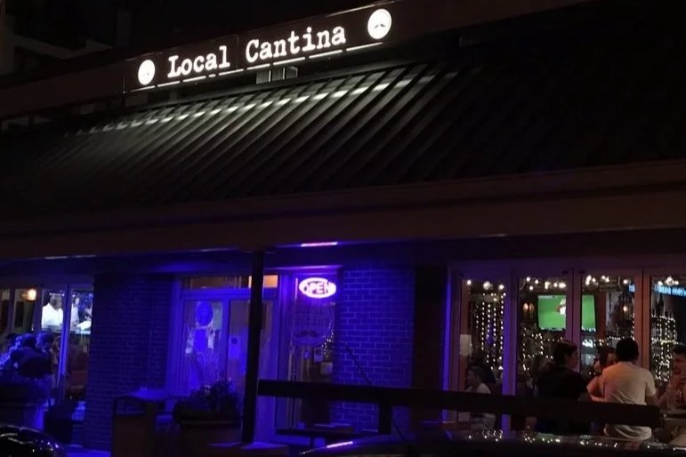Nighttime exterior of a restaurant called 'Local Cantina' with a neon 'Open' sign and people dining inside.