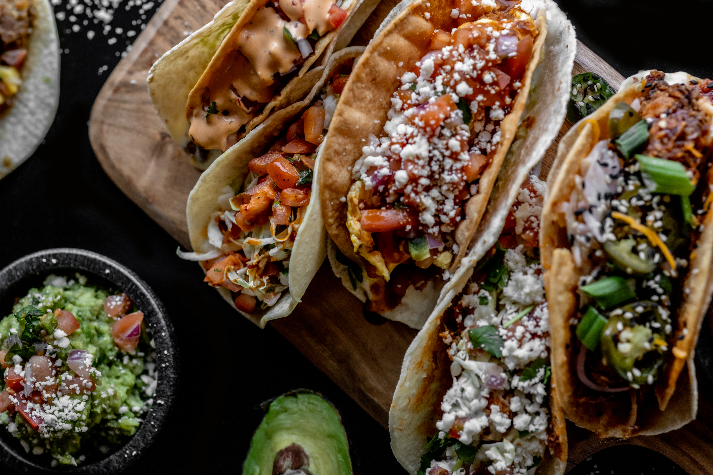 Close-up of five tacos filled with ingredients like diced tomatoes, onions, cilantro, cheese, and sauces, placed on a wooden board with a bowl of guacamole and a halved avocado nearby.