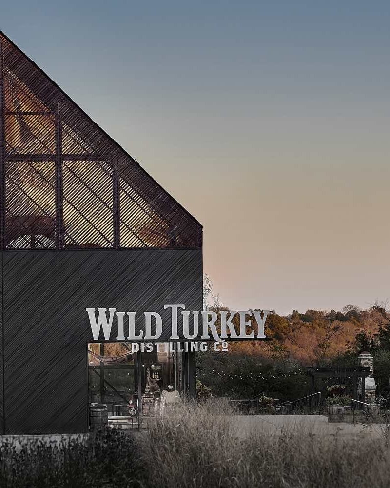 Building with a sign that reads 'Wild Turkey Distilling Co' at sunset, surrounded by trees and tall grass.