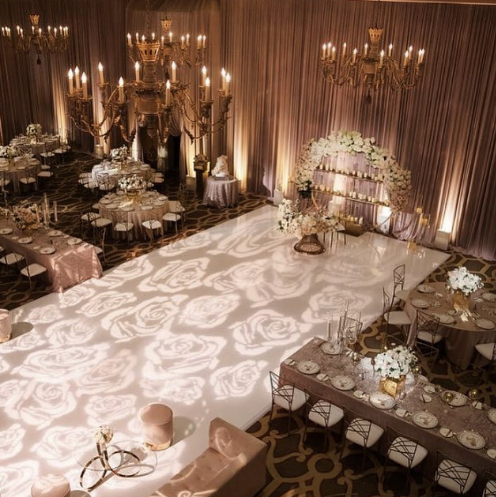 Elegant event space decorated with chandeliers, floral arrangements, and a dance floor with a rose pattern projection. Round tables with floral centerpieces and gold accents create a sophisticated atmosphere for a celebration or wedding.