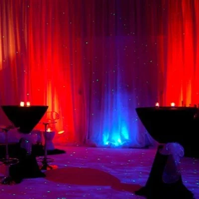 Decorative event setting with red and blue lighting, two cocktail tables with candles, and a blue-lit backdrop.