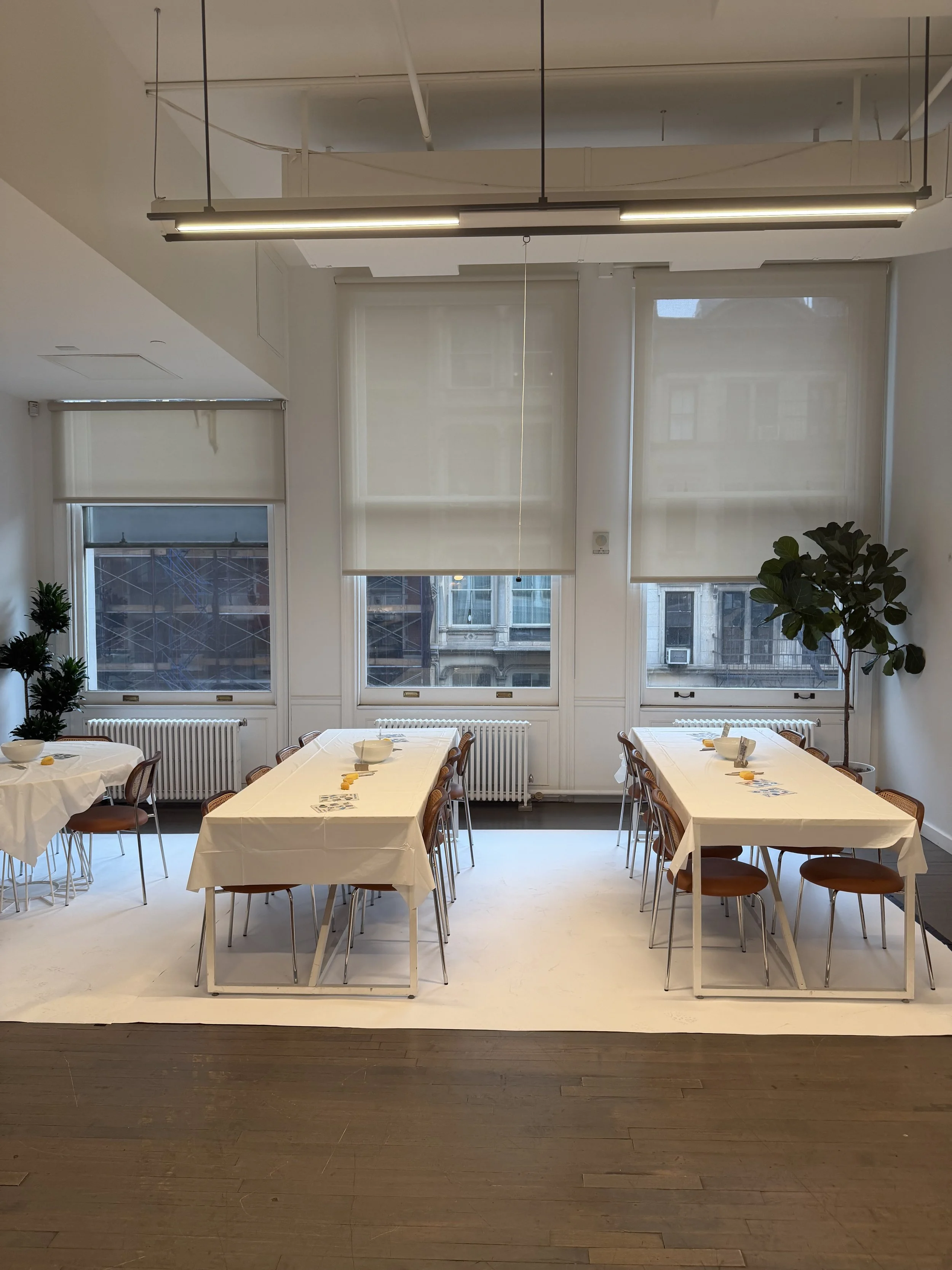 A dining room with white tables and wooden chairs, set with bowls and napkins, large windows with white blinds, and indoor plants, with a wooden floor.