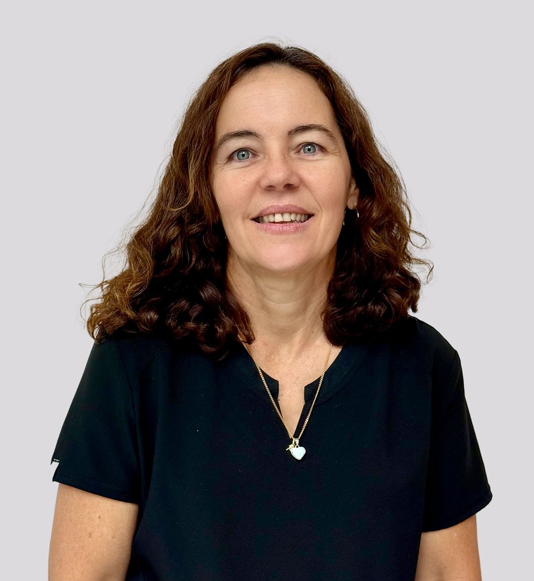 A woman with curly brown hair wearing a black top and a necklace with a heart pendant, smiling against a plain gray background.