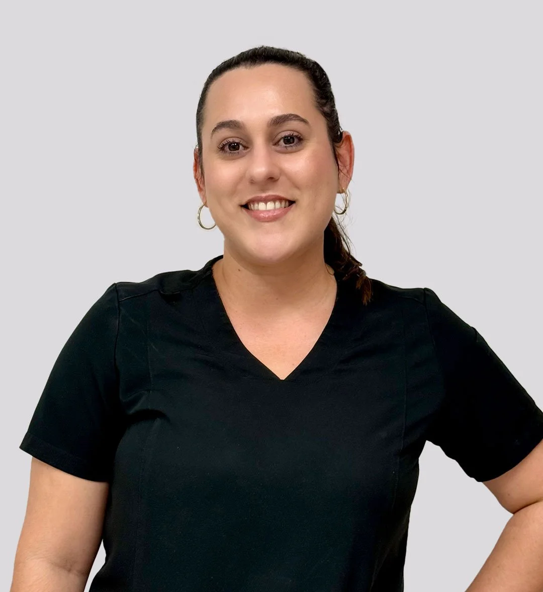 A woman with dark hair tied back, wearing a black short-sleeve top, smiling, with hoop earrings and a neutral gray background.
