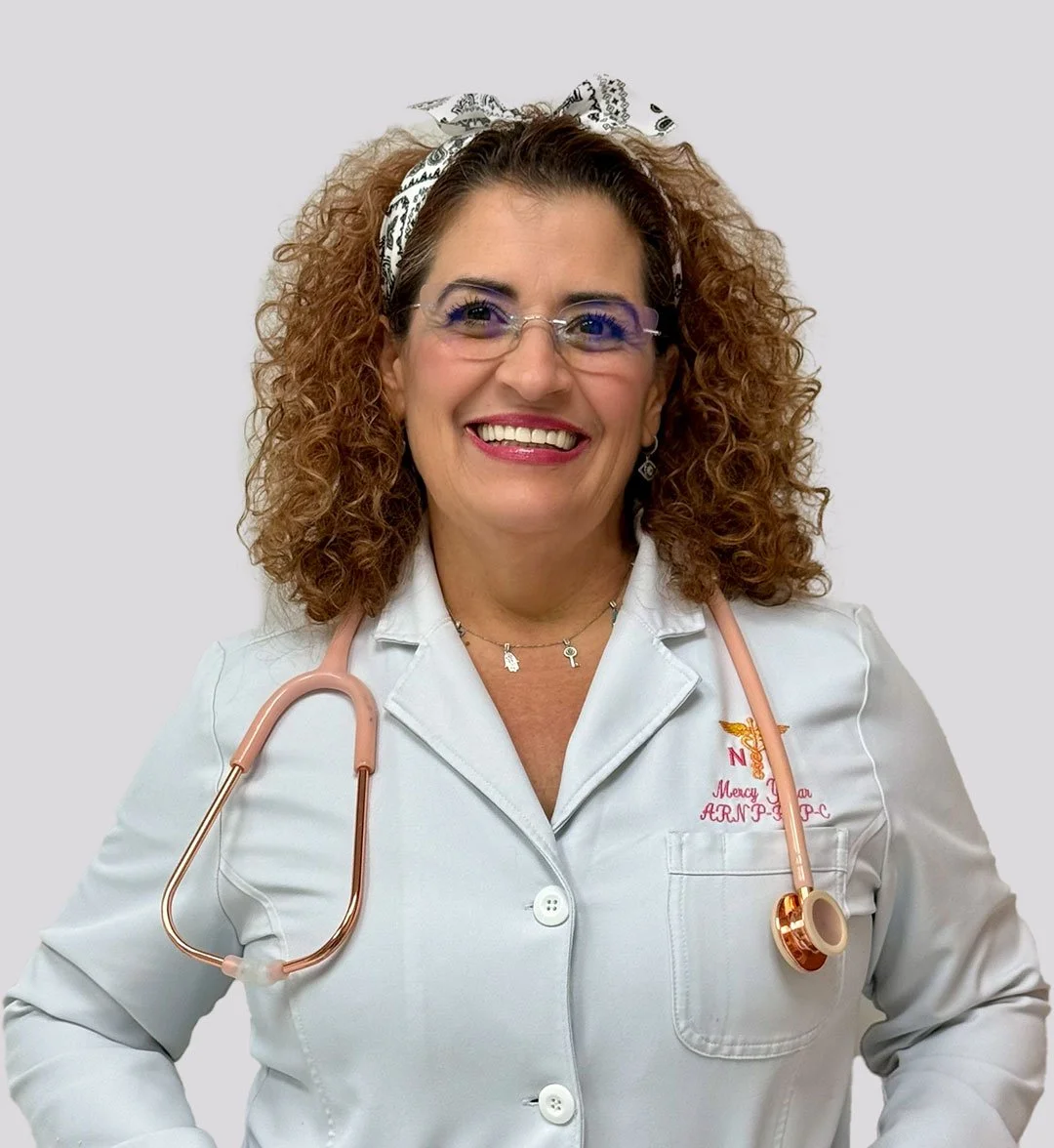 A smiling woman wearing glasses, a white medical coat, and a pink stethoscope around her neck. She has curly hair, a headband, and is standing against a plain background.
