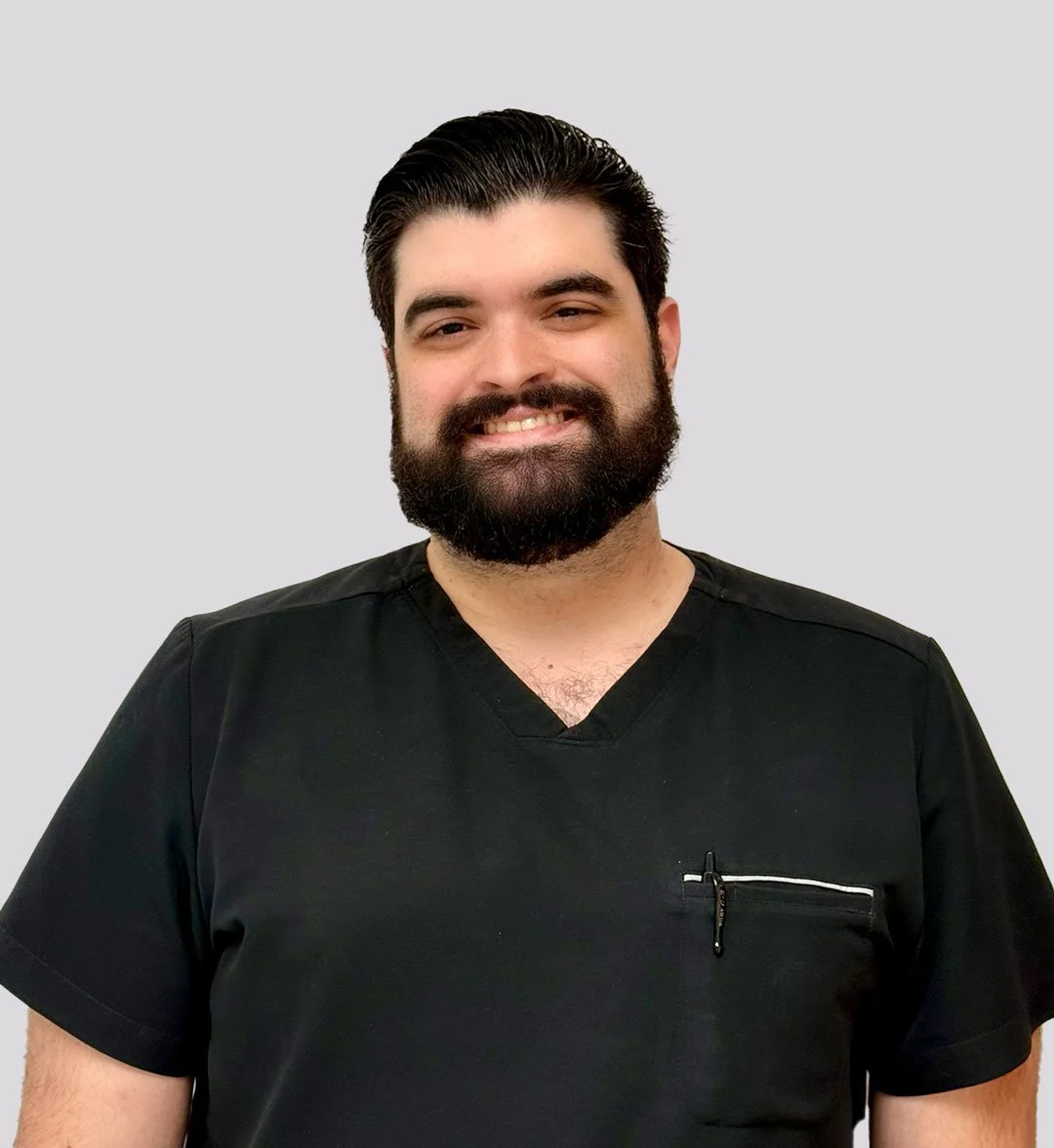 A man with dark hair and a beard, smiling, wearing a black medical scrub top, standing against a plain light gray background.