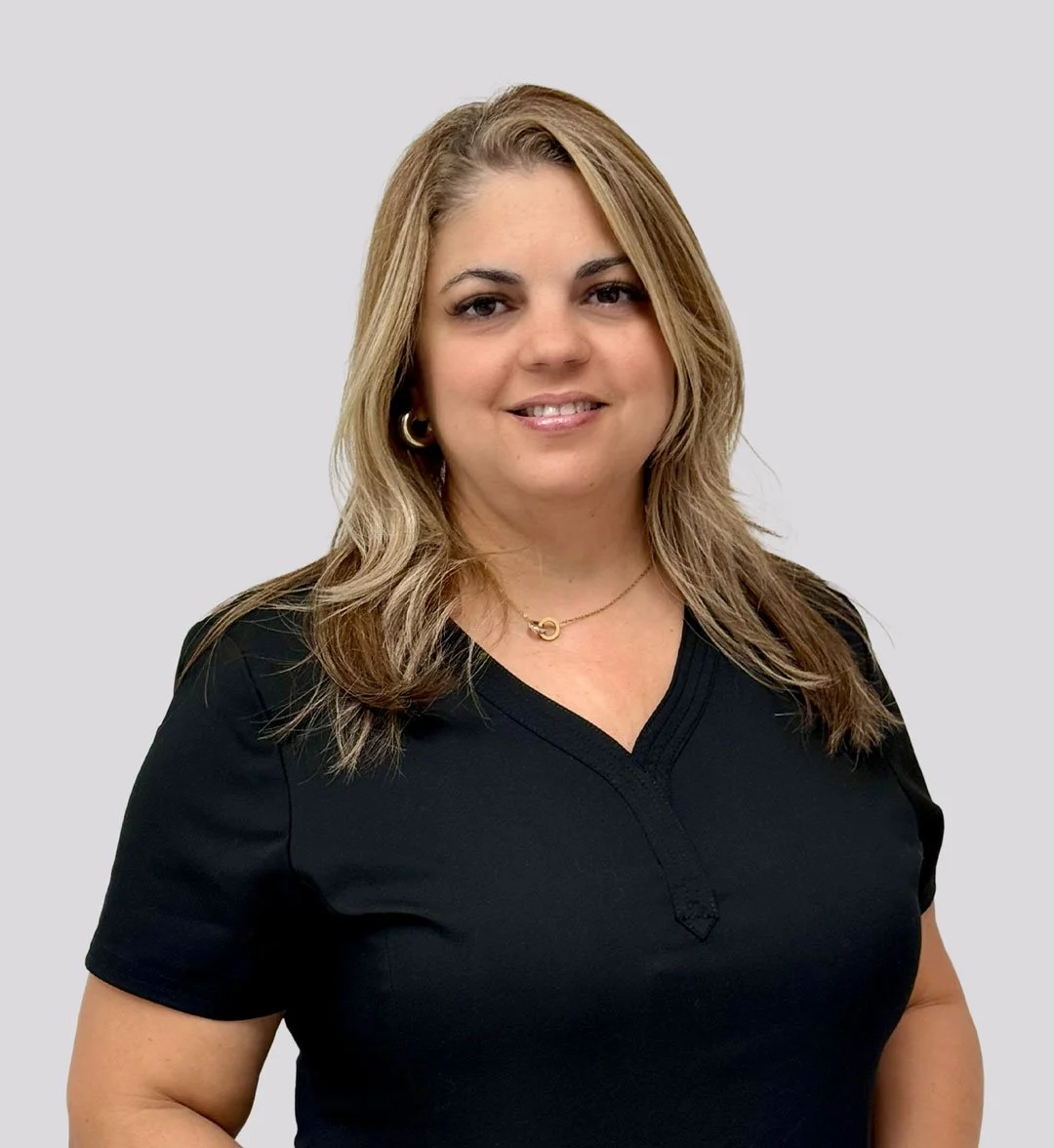 A woman with shoulder-length light brown hair, wearing a black V-neck top, gold hoop earrings, and a gold necklace, standing against a plain light gray background.