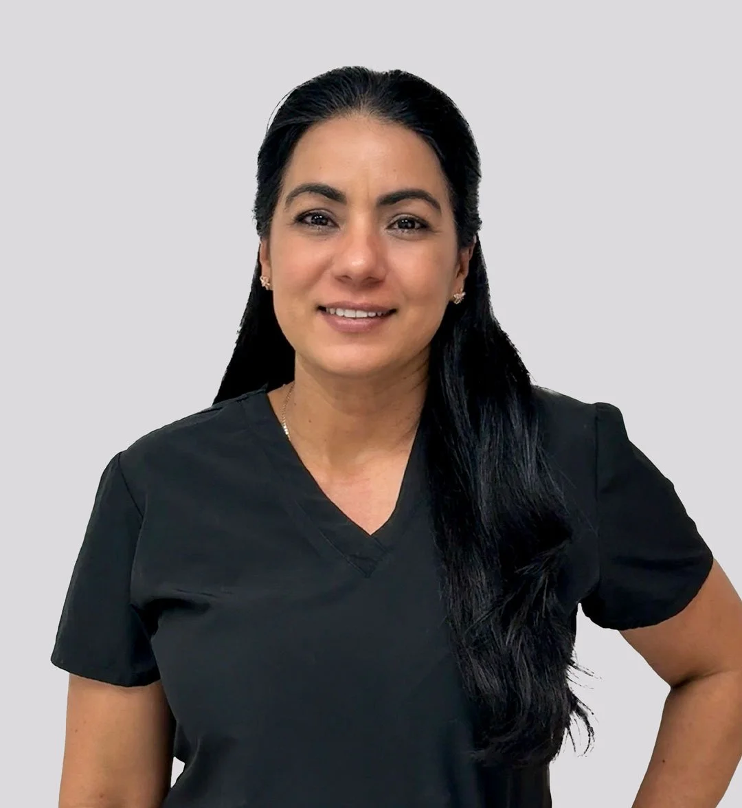 A woman with long black hair, wearing a black V-neck shirt, smiling against a plain light gray background.