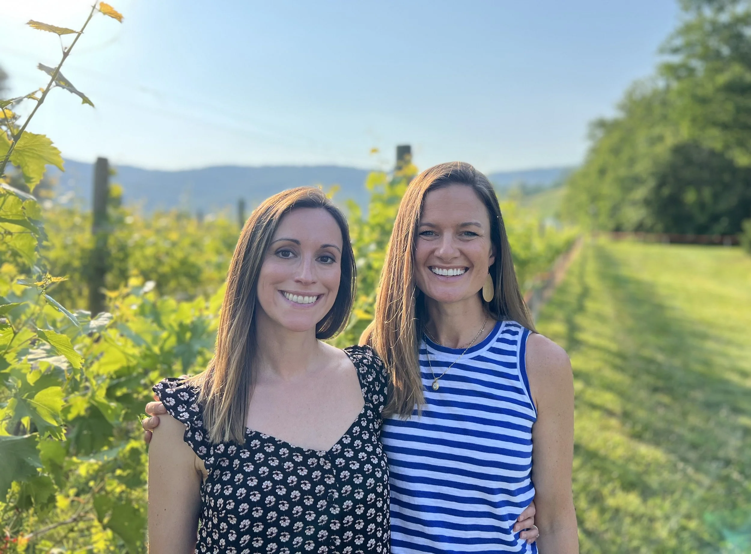 Katelin and Kristen standing outdoors in a vineyard with green plants and mountains in the background.
