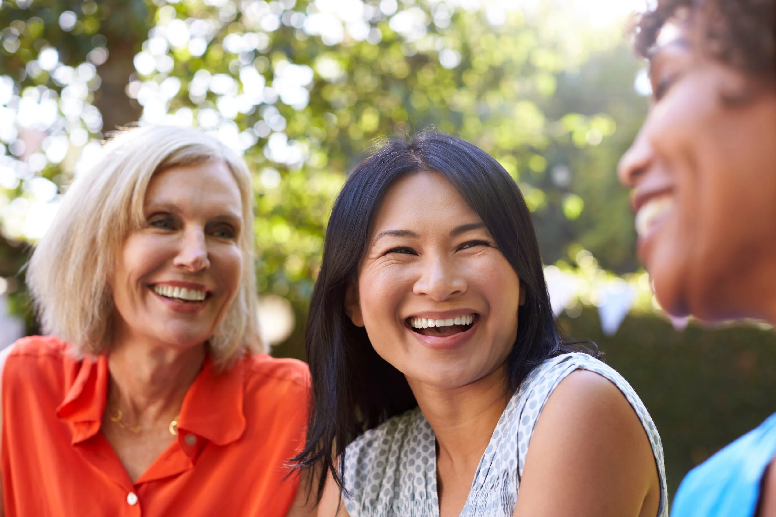 A group of three women smiling outdoors with a blurred background of trees and sunlight.