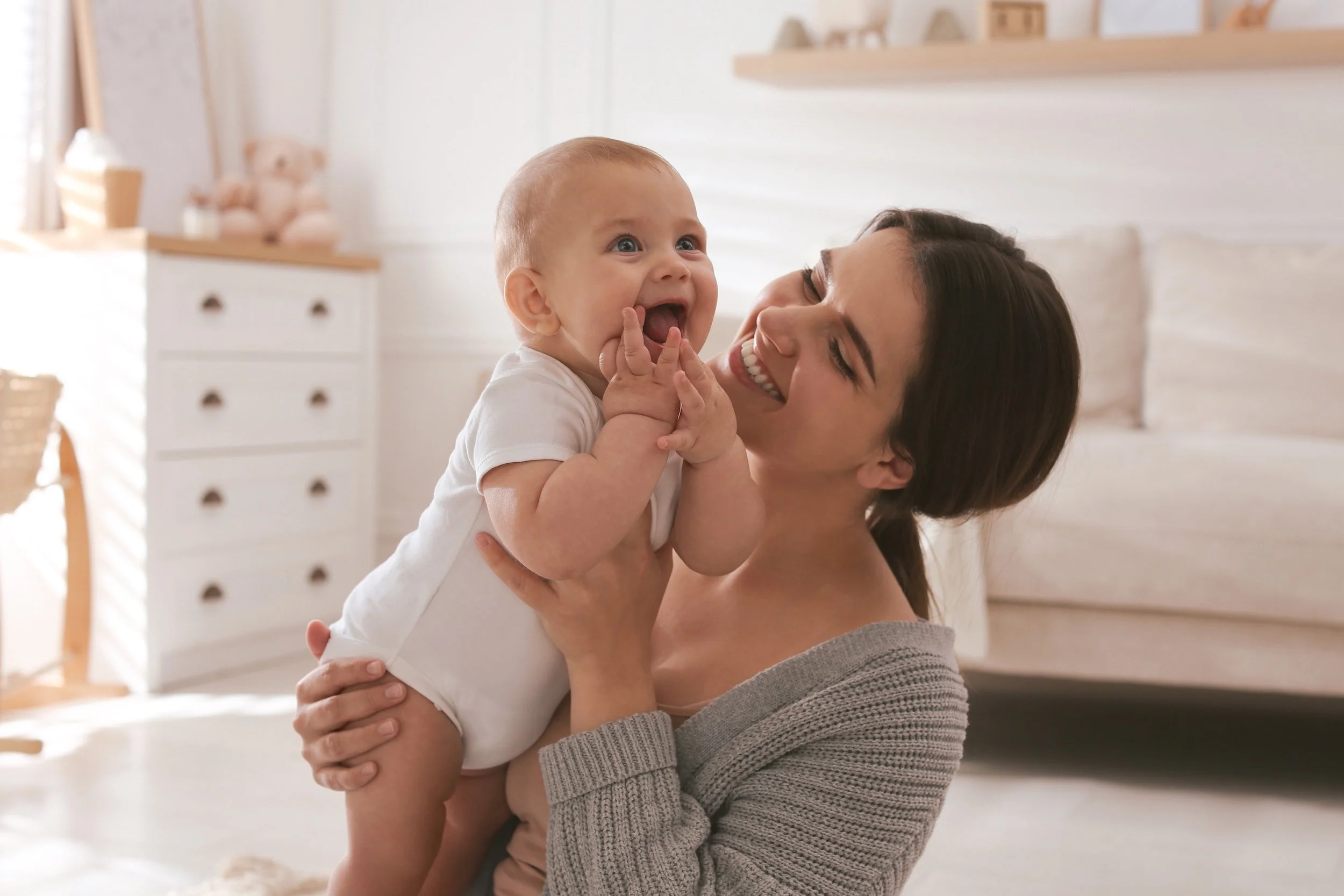 A woman with dark hair smiling and holding a happy baby with light hair and blue eyes in a bright, cozy bedroom.