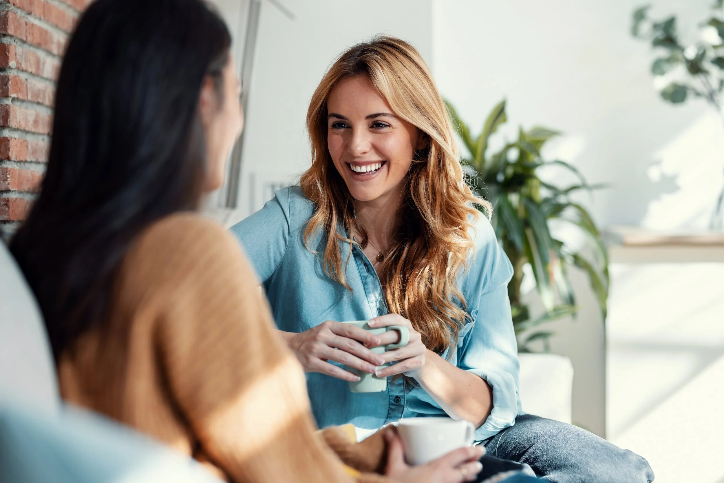 Two women having a conversation in a bright, modern room with plants. One woman with long dark hair is partially visible, and the other with wavy red hair is smiling while holding a mug.