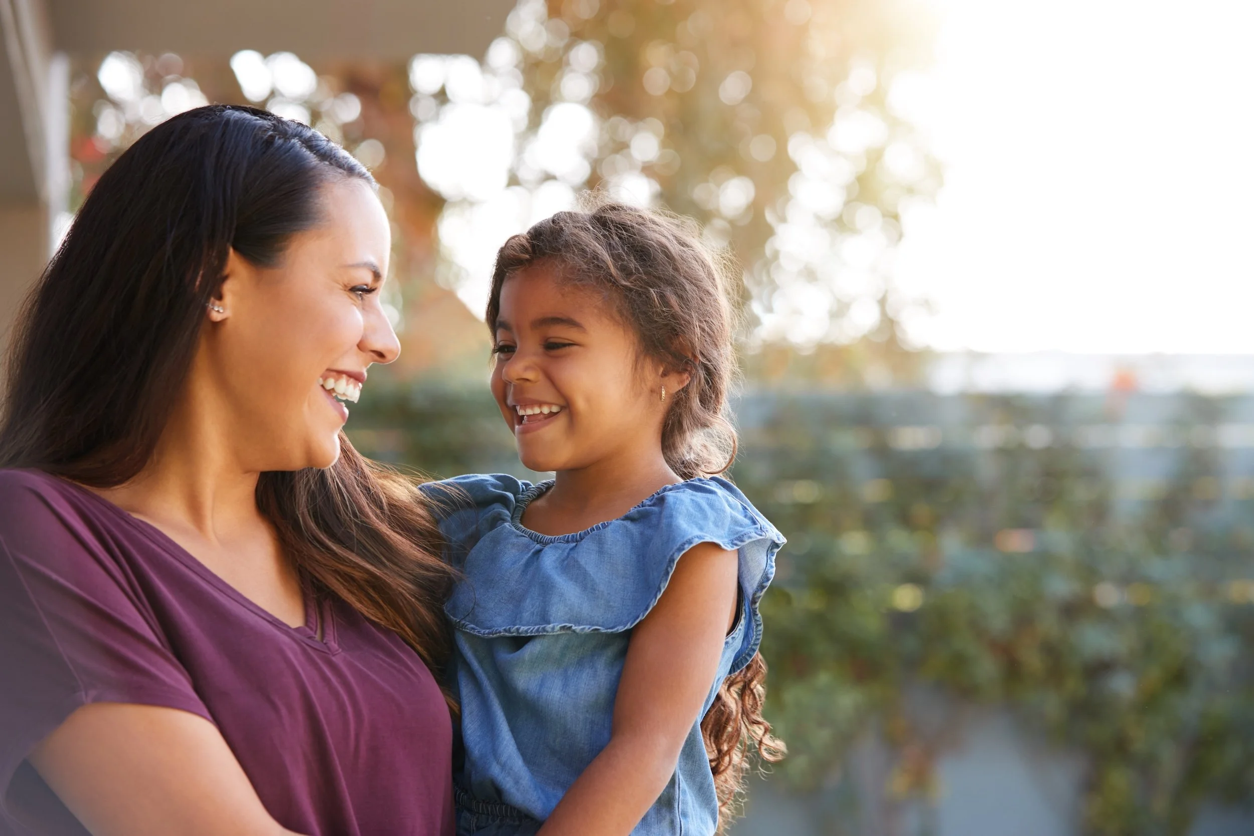 A woman with dark hair and earrings smiling at a young girl with light brown hair, both outdoors with trees in the background. The woman wears a purple top and the girl wears a blue dress.