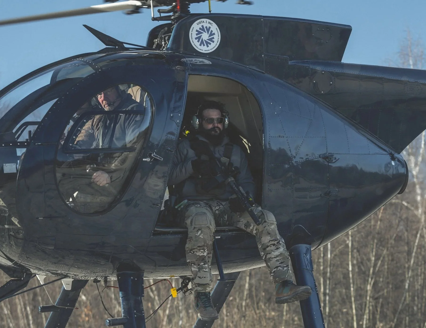 A military-style helicopter with two pilots inside, one sitting near the open door and the other in the cockpit, both wearing tactical gear and sunglasses, flying over a wooded area.