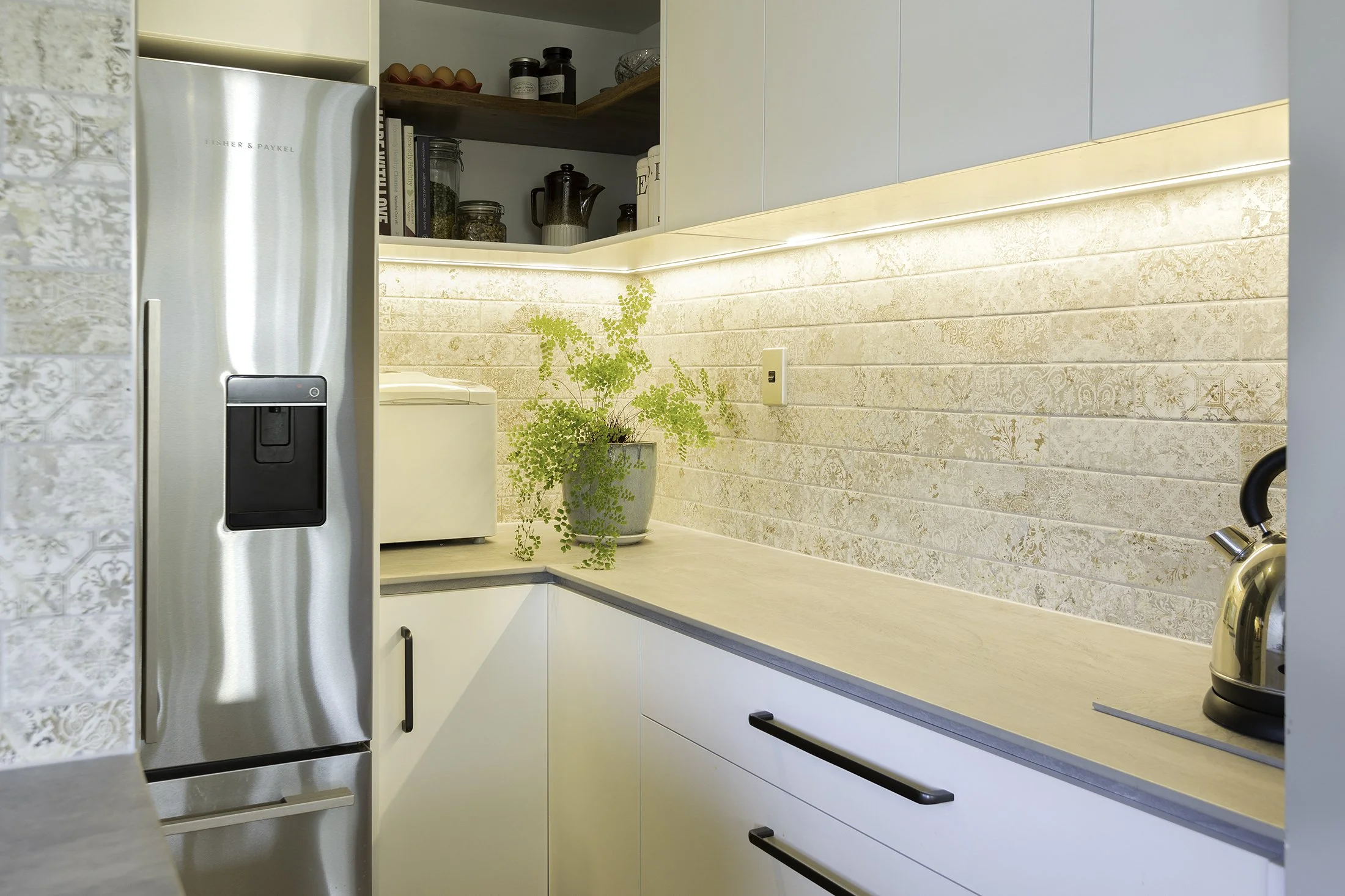 Kitchen corner with a stainless steel refrigerator, a potted green plant, a toaster, and a kettle on a beige countertop with a beige textured backsplash. Open shelves above hold books, jars, and kitchenware.