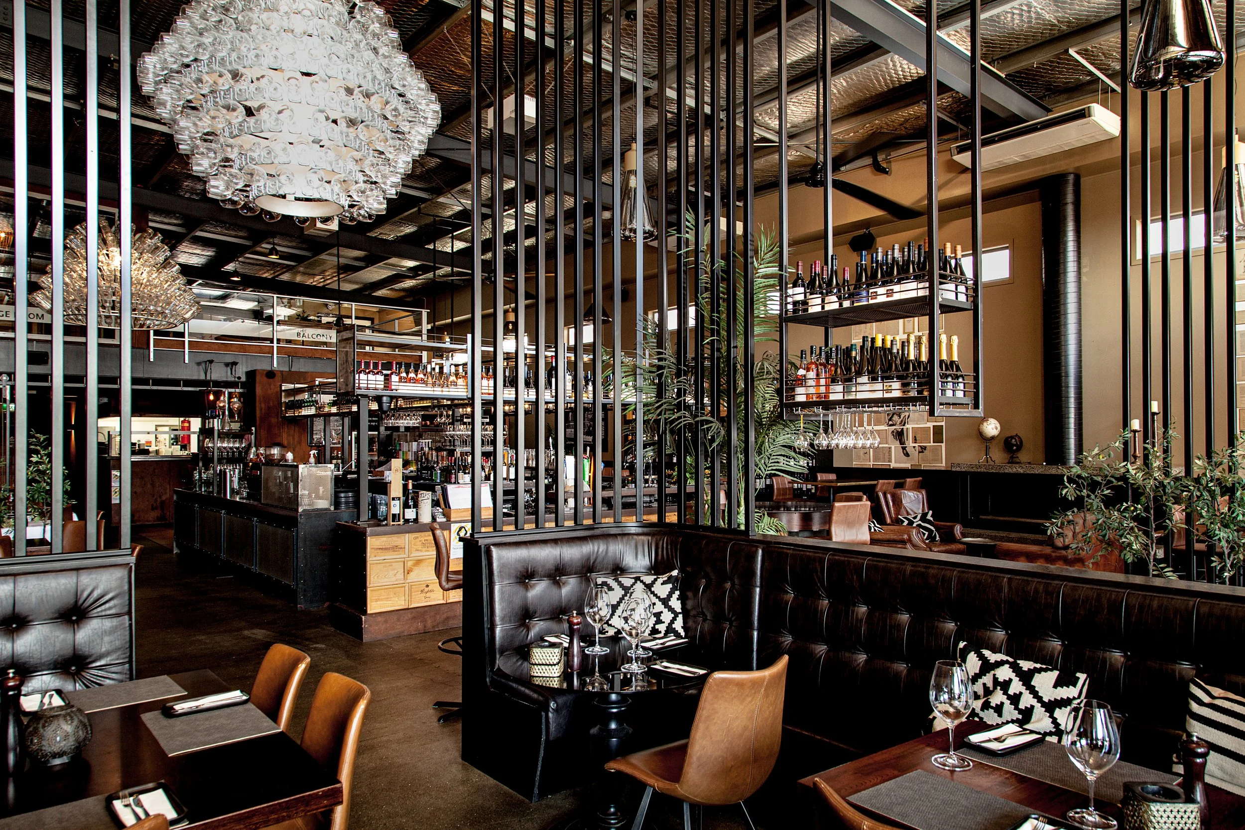 Interior of a stylish restaurant or bar with modern decor, including wooden furniture, leather seating, a chandelier, and a bar area with bottles and glasses.