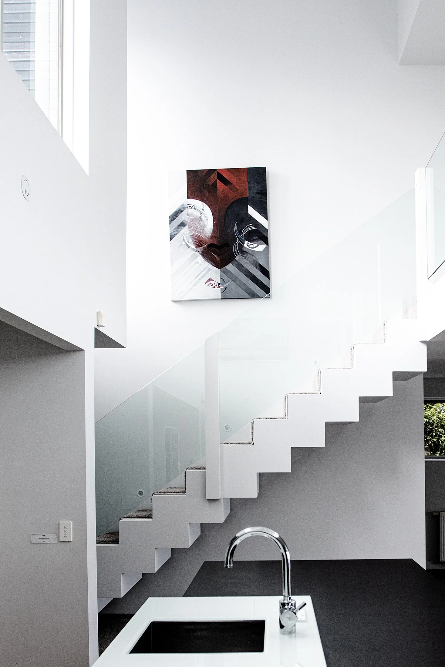 Modern minimalist interior of a white staircase with glass railing, a black and white abstract face painting on the wall, and a kitchen island with a sink in the foreground.
