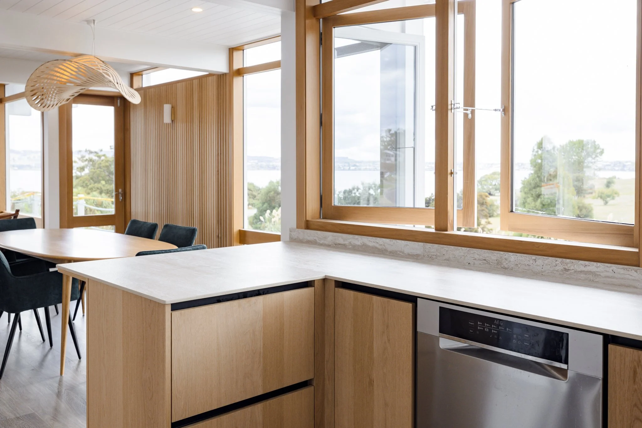 Modern kitchen and dining area with large windows, wooden accents, and a view of trees and a body of water outside.