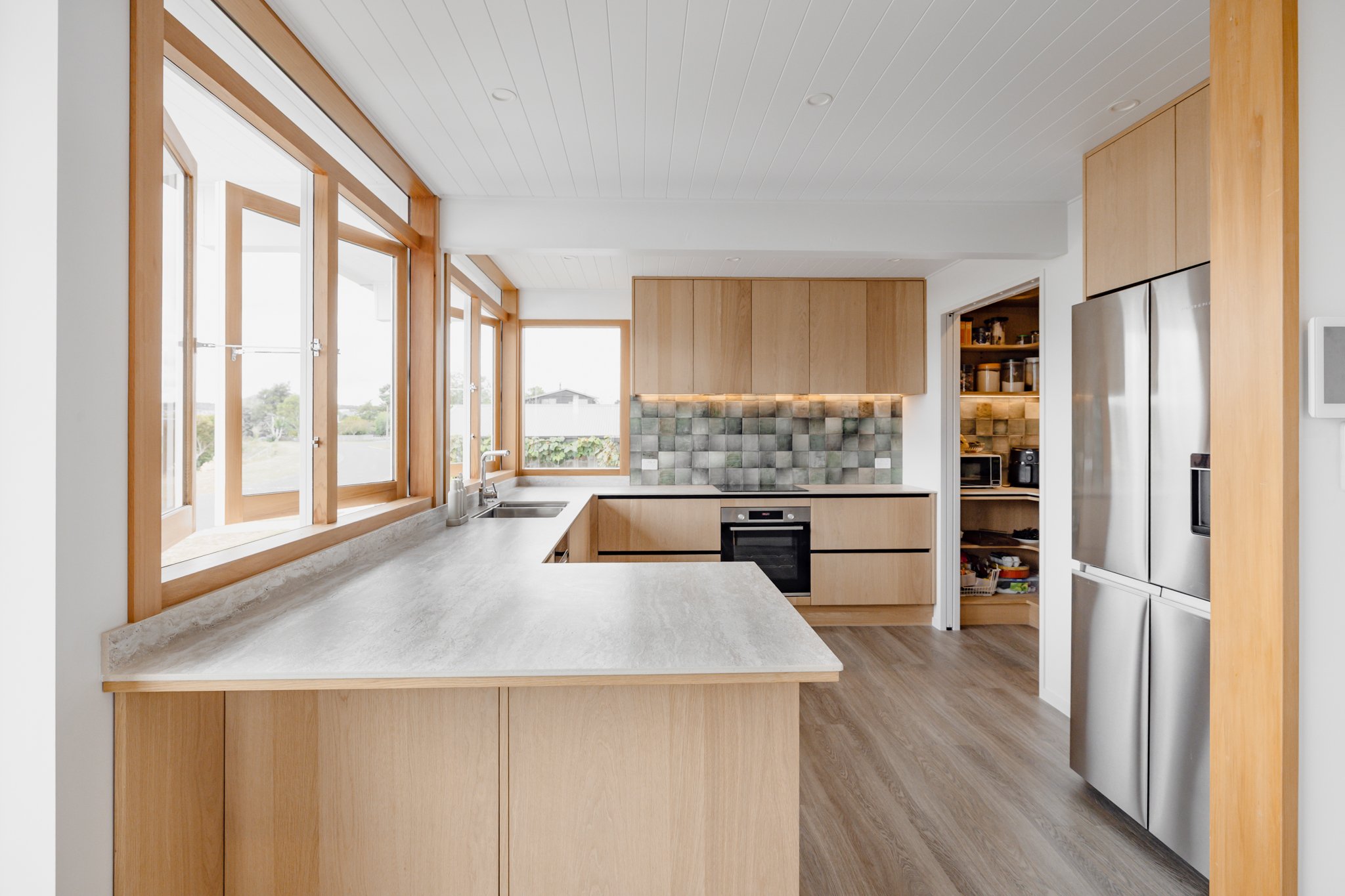 Modern kitchen with large window, light wood cabinets, gray countertop, white ceiling with recessed lighting, backsplash with gray tiles, built-in oven, and open pantry with shelves.