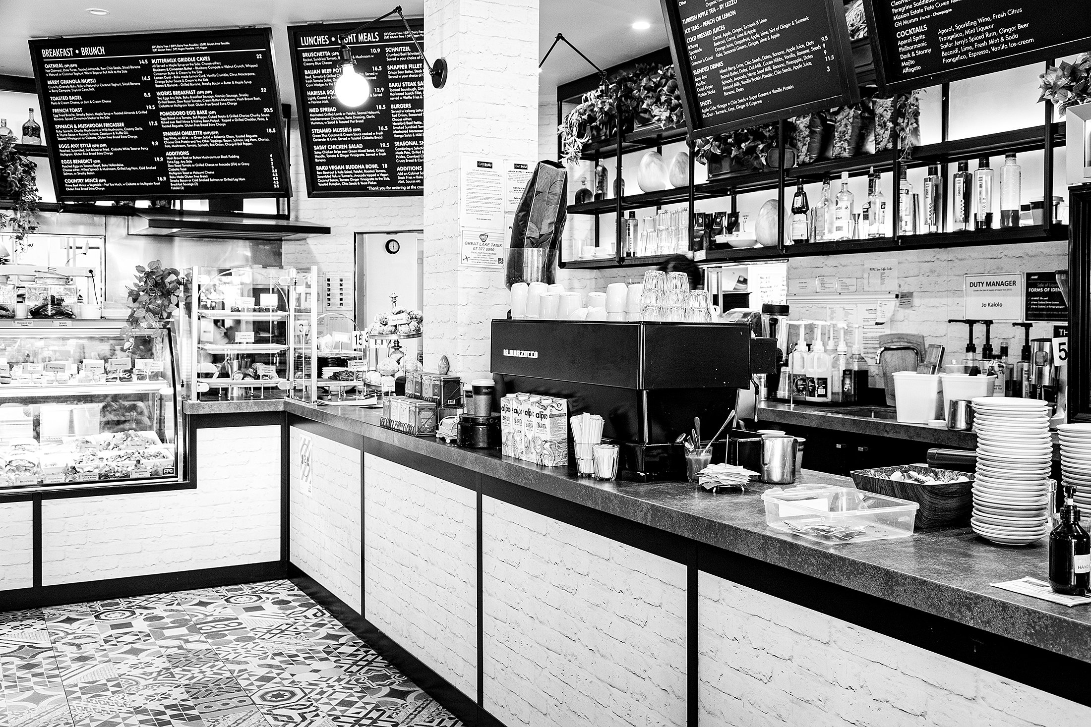 Black and white photo of a cafe counter with menu boards on the wall, coffee machine, stacks of plates, glassware, and various supplies on the countertop, with patterned floor tiles.