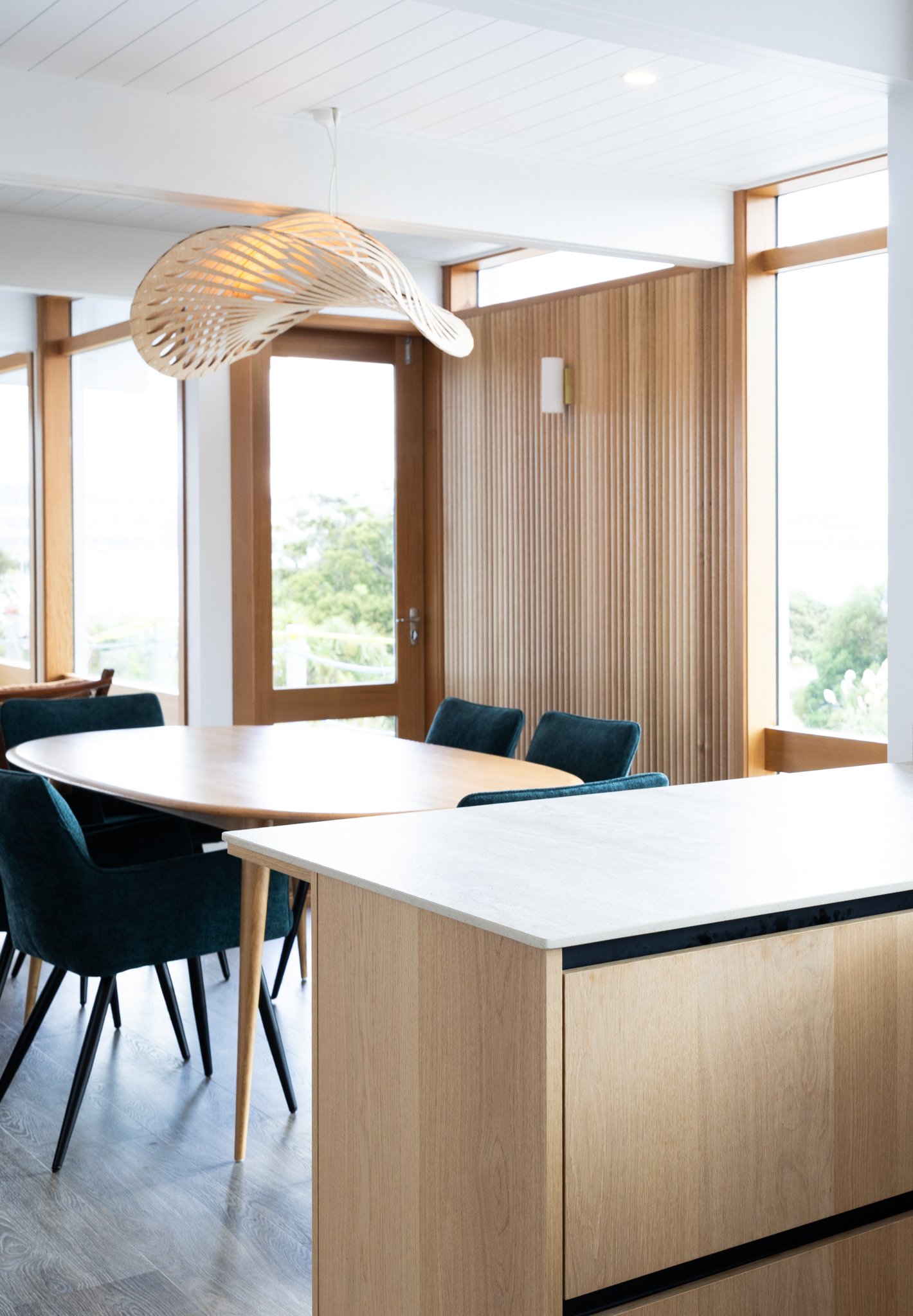 A modern dining area with a wooden table, dark upholstered chairs, large windows, light wood paneling, and a unique wooden pendant light fixture.