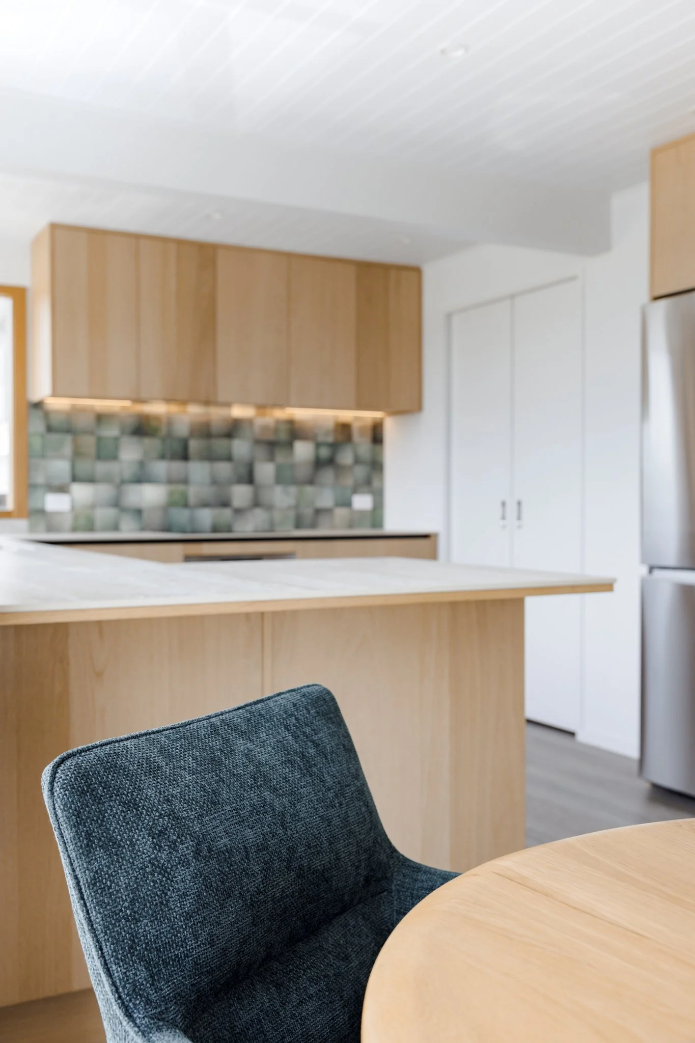Modern kitchen with wood cabinets, a mosaic tile backsplash, and a light-colored countertop, seen from a dining area with a dark gray upholstered chair.