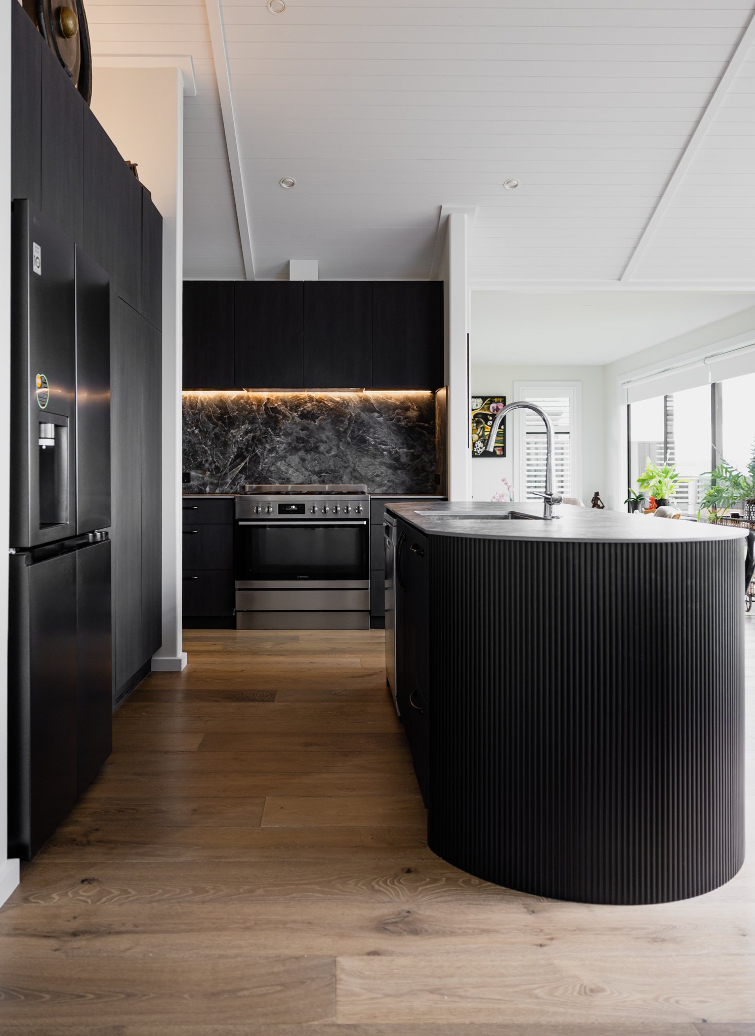 Modern kitchen with black cabinetry, marble backsplash, stainless steel appliances, and a curved black island with a sink, alongside hardwood flooring and large windows.