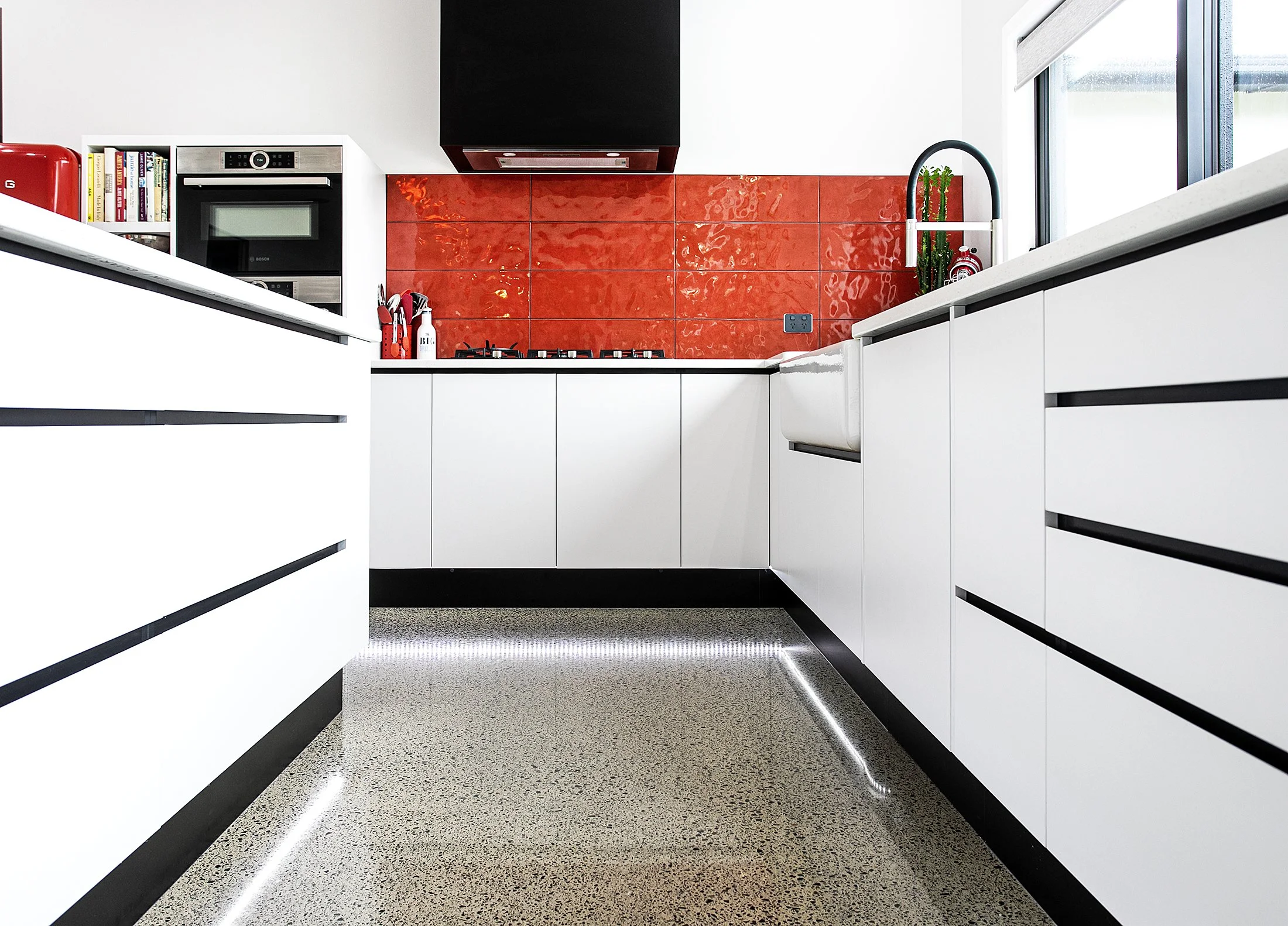 Modern kitchen with white cabinets, red tile backsplash, black accents, and a large window with roller blinds.