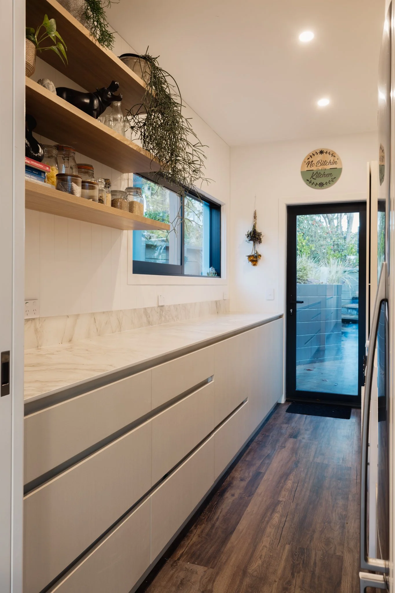 Modern kitchen with white cabinetry, marble countertops, open wooden shelves with jars and plants, a large window, and a glass door leading outside.