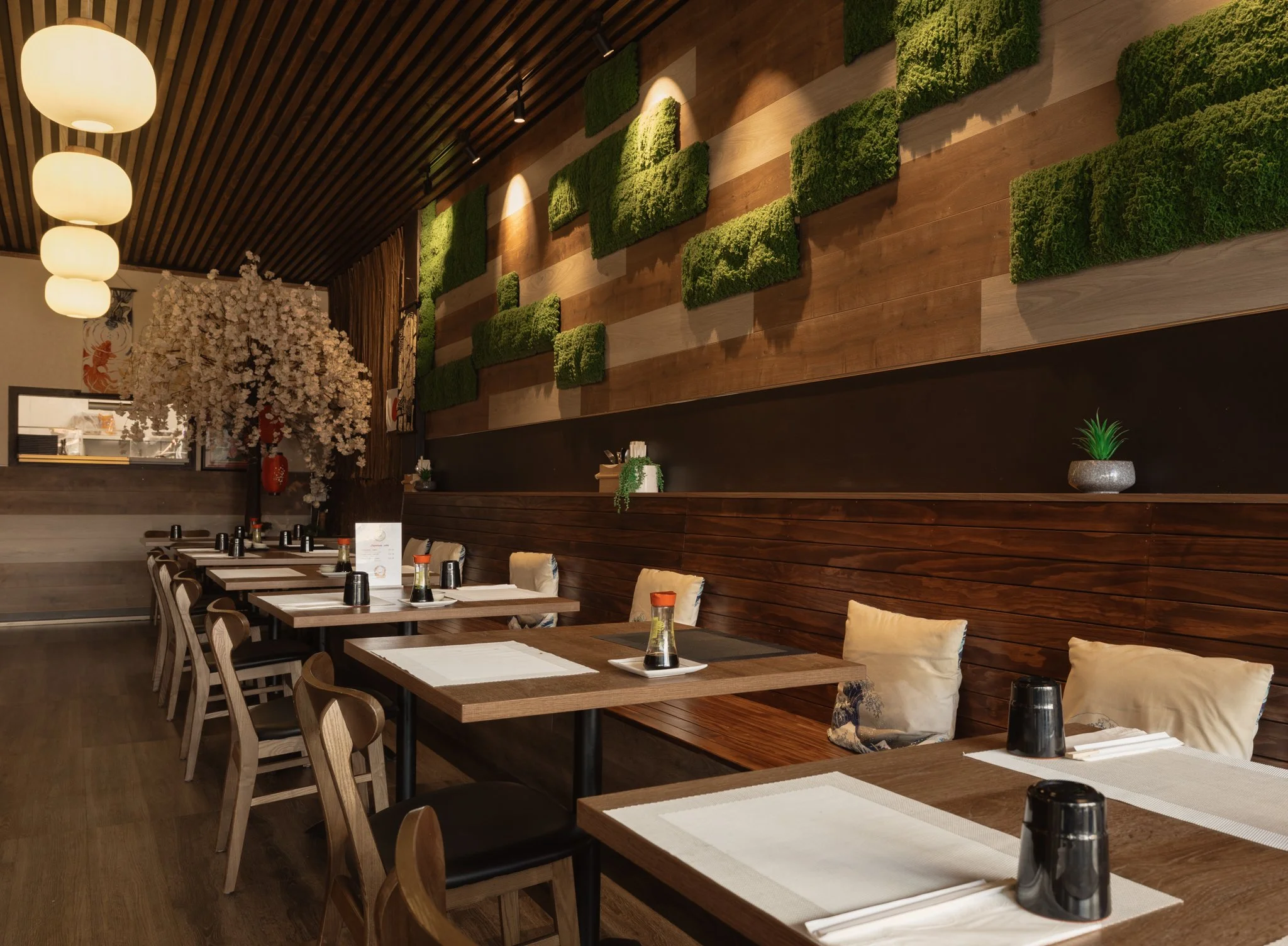 Interior of a restaurant with wooden tables and chairs, decorative wall with green moss art, hanging paper lanterns, and cherry blossom decor.