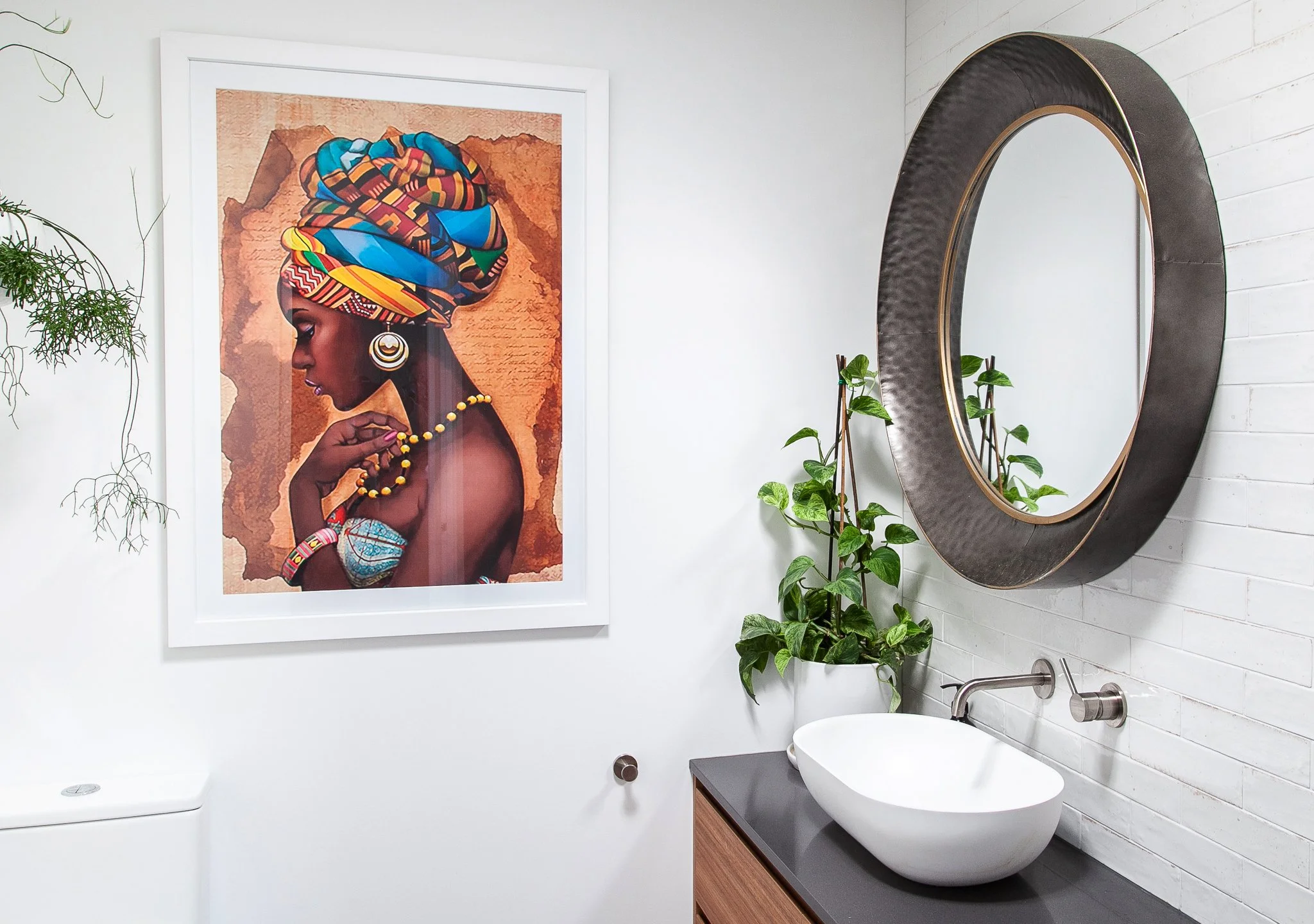Modern bathroom with a large circular mirror, plants, and a framed artwork of a woman in traditional African attire on the wall.