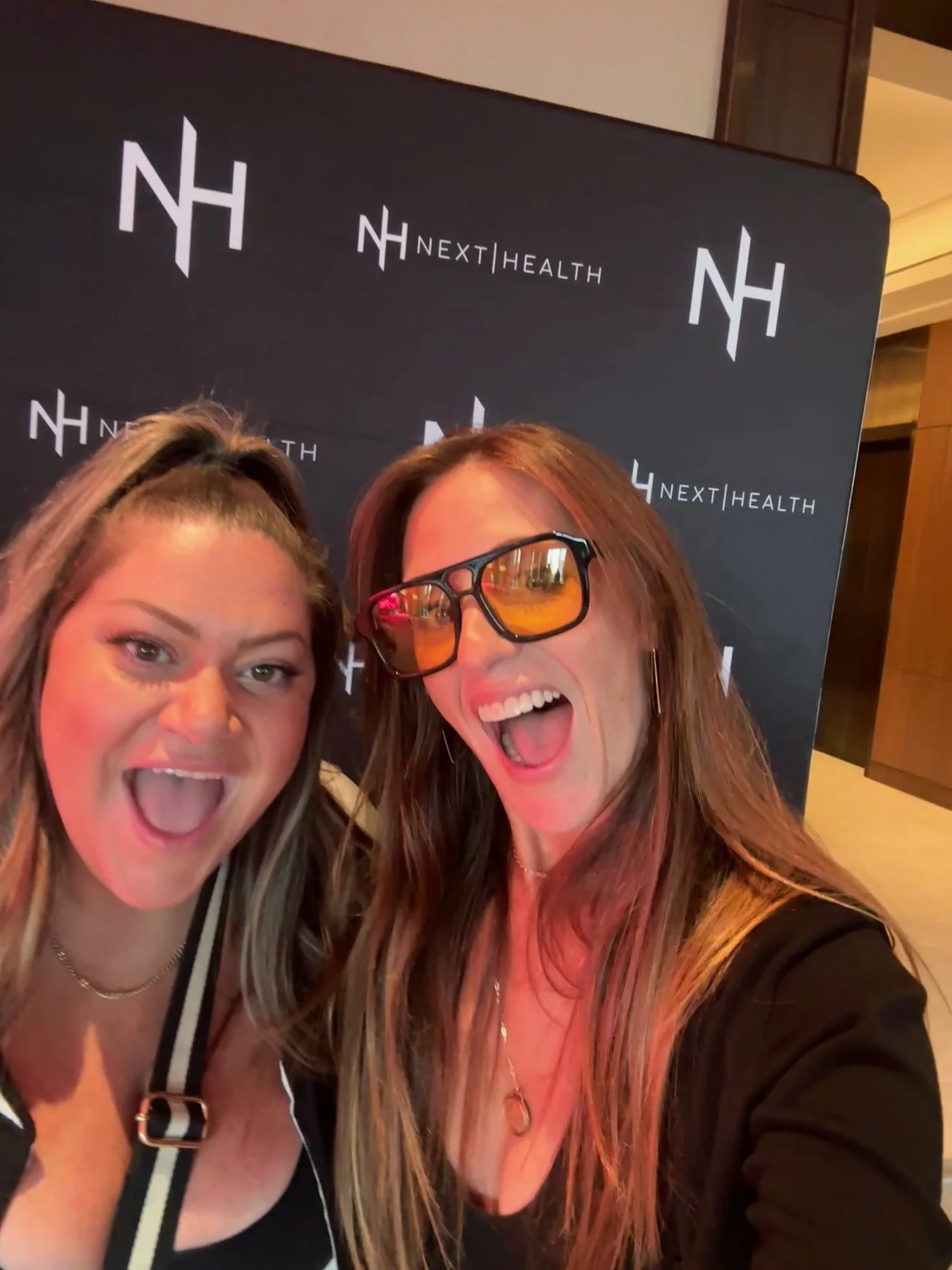 Two women taking a selfie in front of a black backdrop with the text and logo 'NextHealth' on it. Both women are smiling with open mouths and wearing glasses.