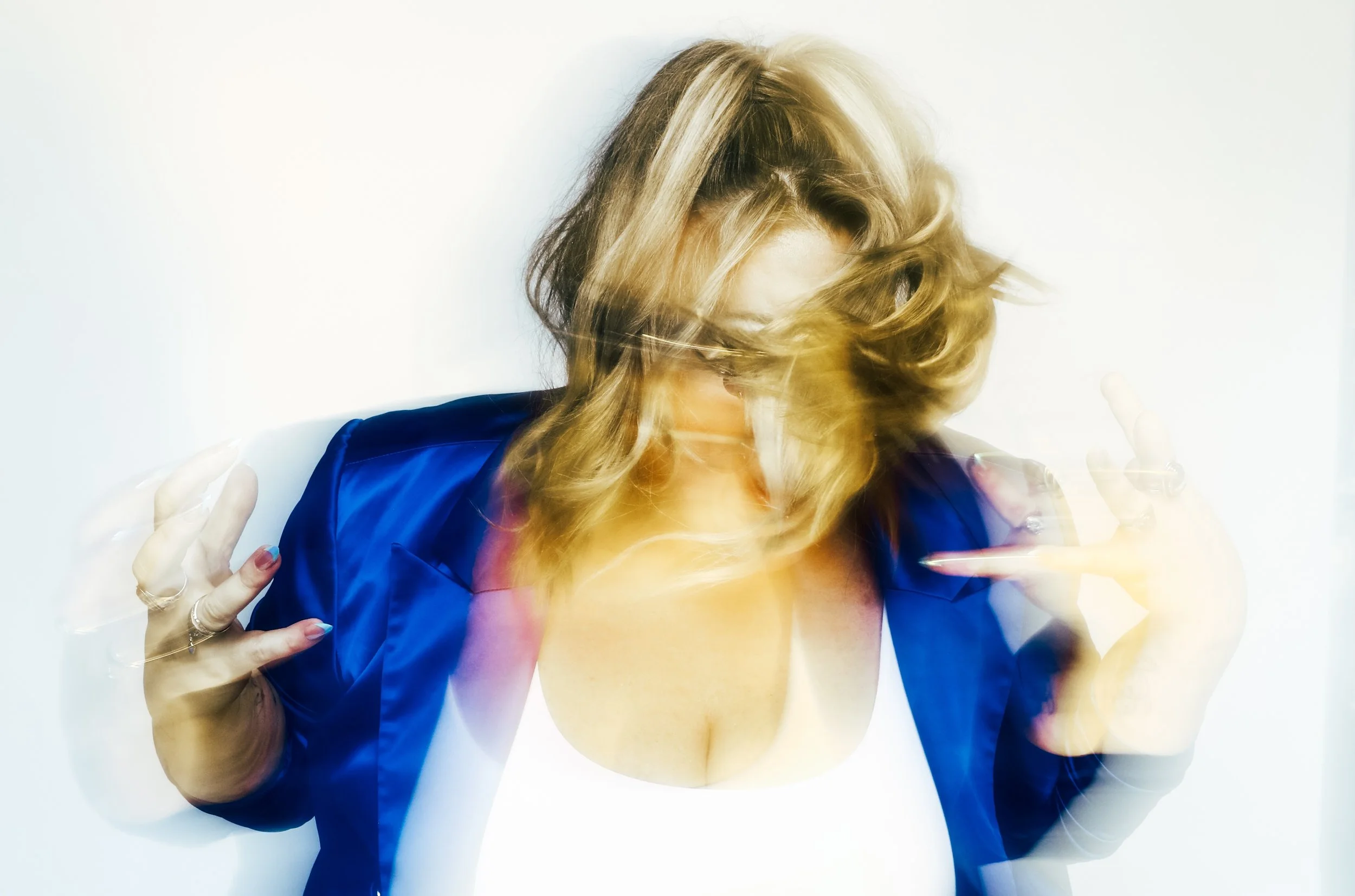 Blonde woman with wavy hair wearing a shiny royal blue jacket, facing forward with her head tilted down and her hair covering her face, hands raised with fingers pointing or making gestures, against a plain white background.