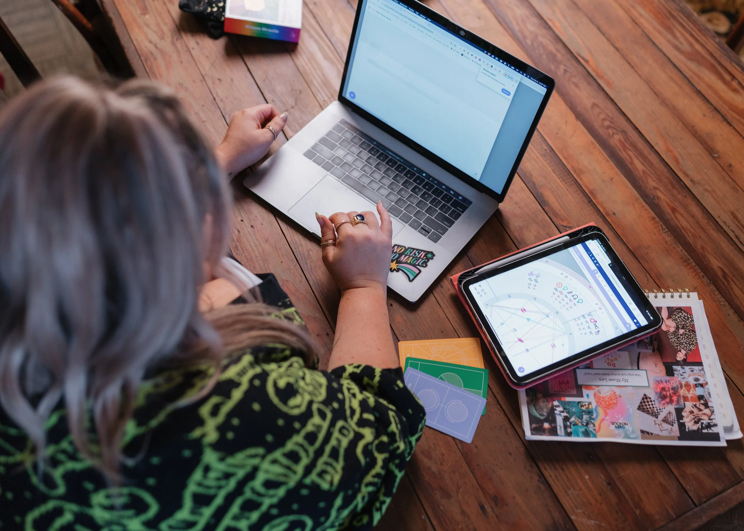A person with long, gray hair sitting at a wooden table, using a silver laptop. The laptop screen displays a messaging app. On the table are various colorful cards, papers, and a tablet showing a colorful diagram or calendar.
