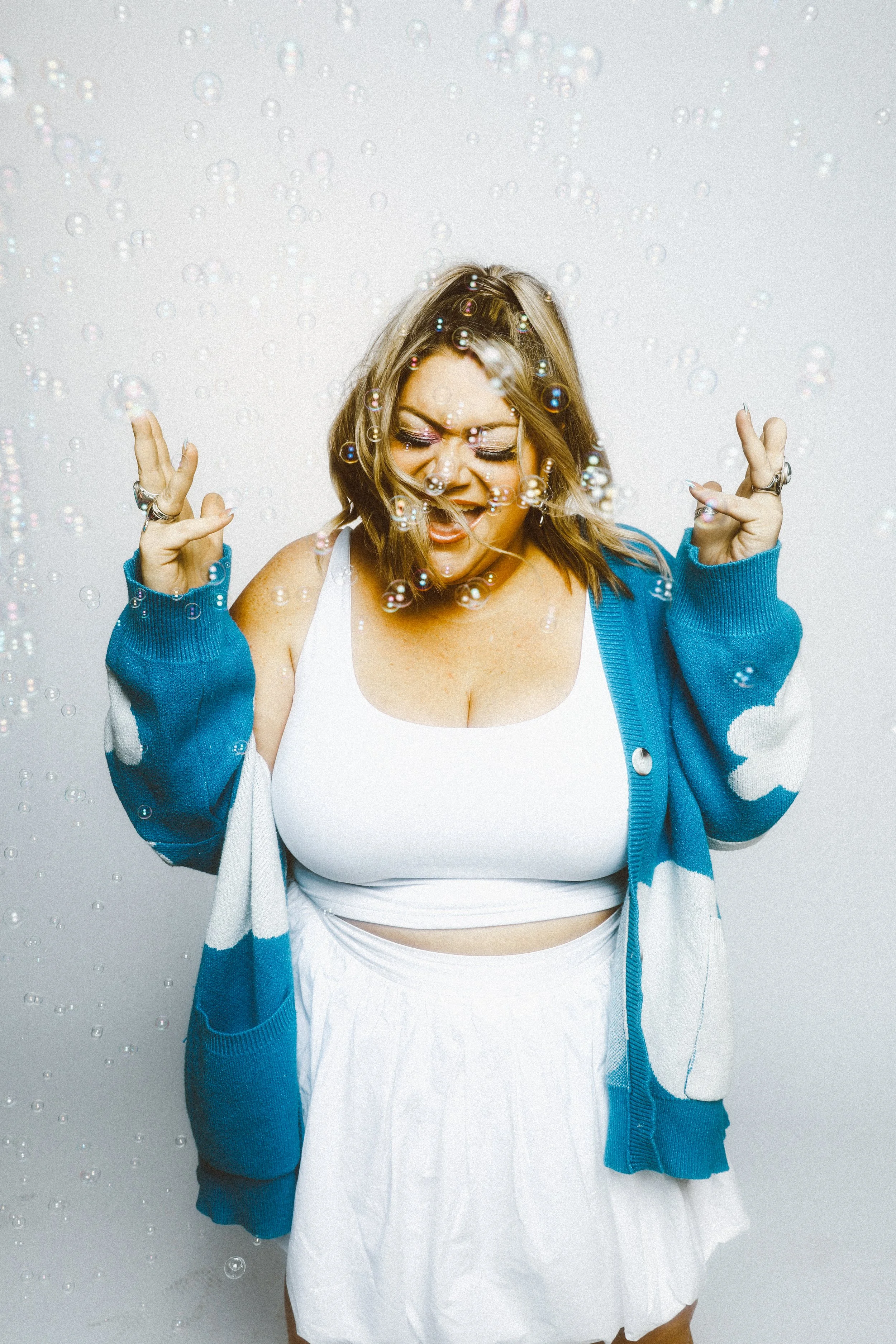 A woman with shoulder-length blonde hair, wearing a white crop top and a blue and white hoodie, stands against a white background with water droplets, holding her hands up in a fun gesture while bubbly soap bubbles surround her.