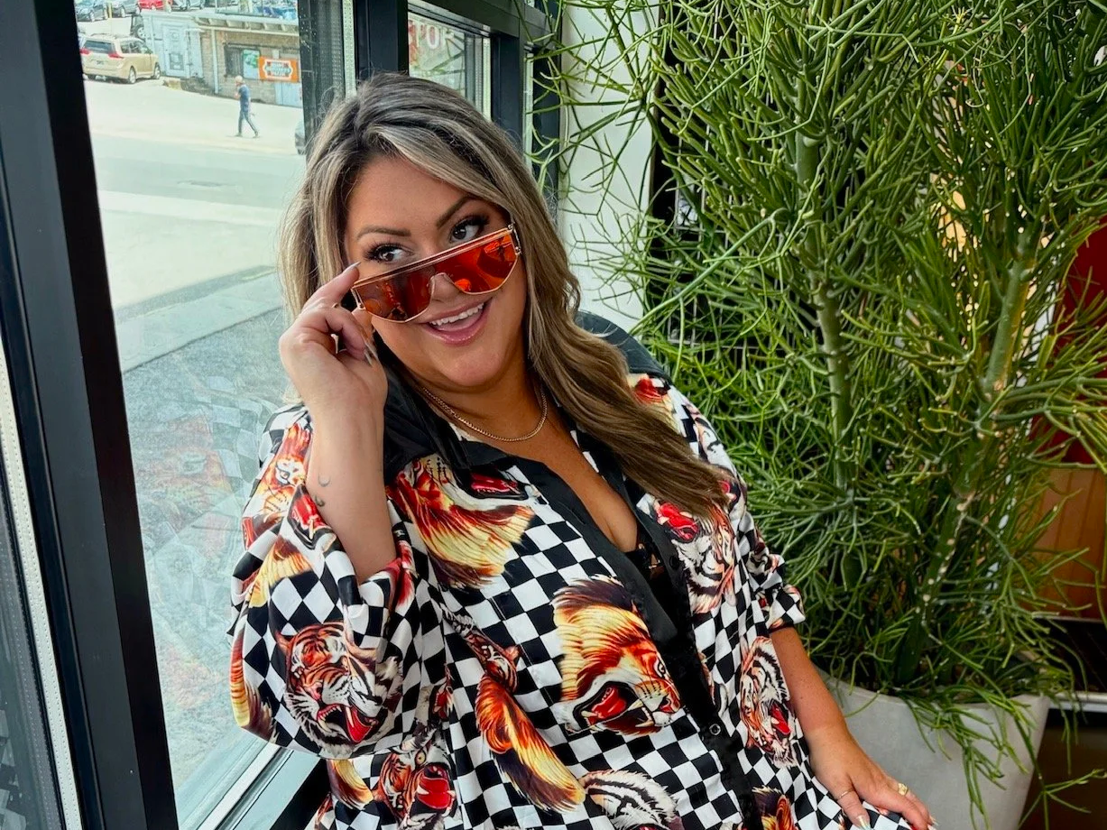 A woman with long brown hair and sunglasses posing indoors next to a large green plant, wearing a black and white checkered shirt with lion faces and tiger images.