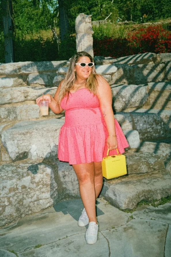 Woman in a pink dress with white shoes and sunglasses, holding a yellow bag in one hand and a drink in the other, standing in front of stone steps and a carved stone owl sculpture outdoors.