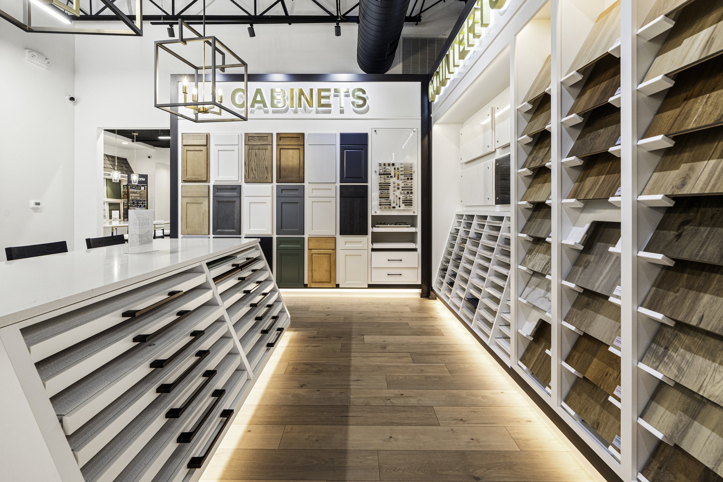 Interior of a showroom with display shelves featuring various cabinet door styles and wood finishes, with a large illuminated sign that reads 'CABINETS' on the back wall.